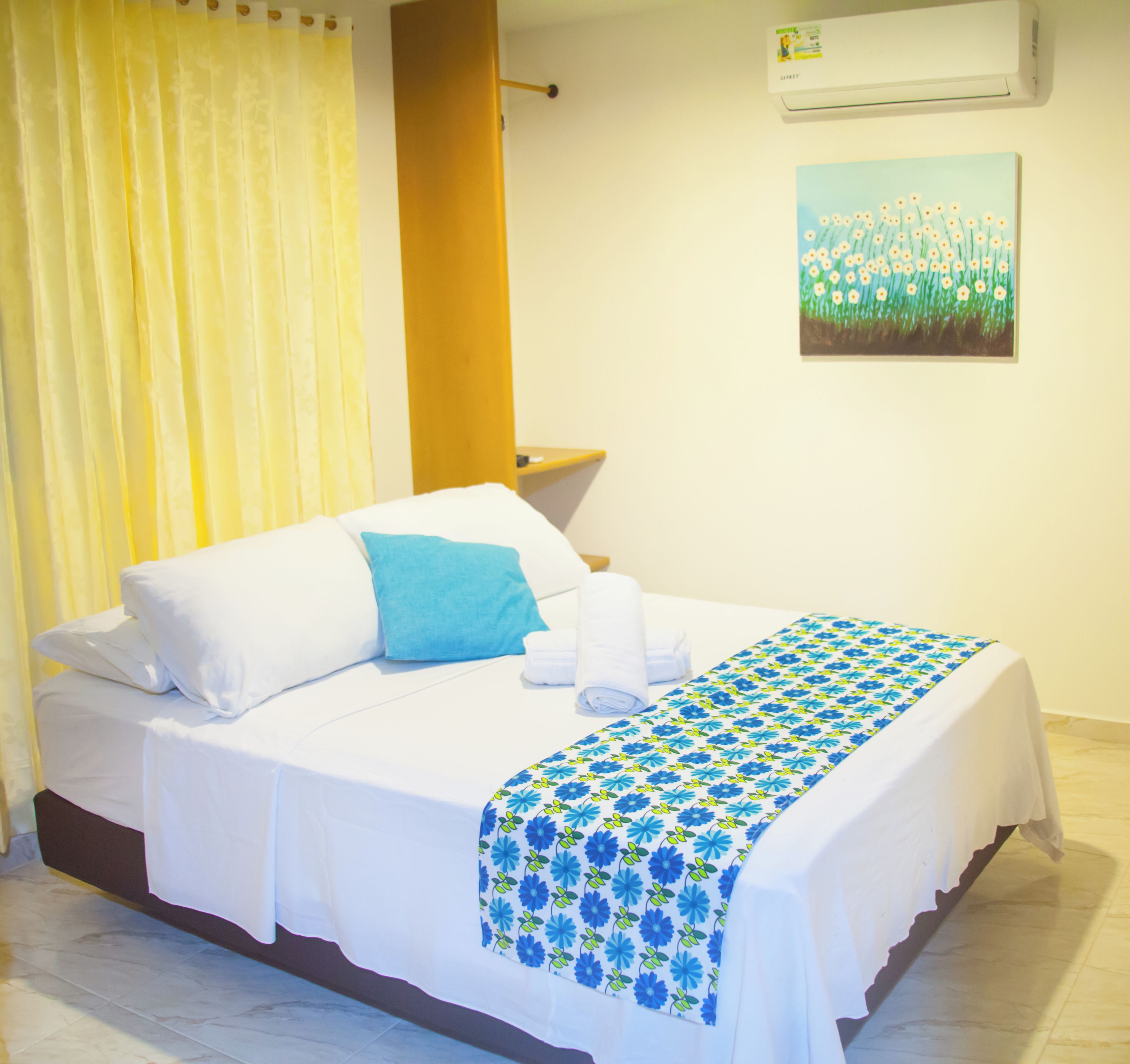 A neatly made bed with white linens and a blue floral bedspread, a yellow curtain, a wooden headboard, and a painting of flowers on the wall.