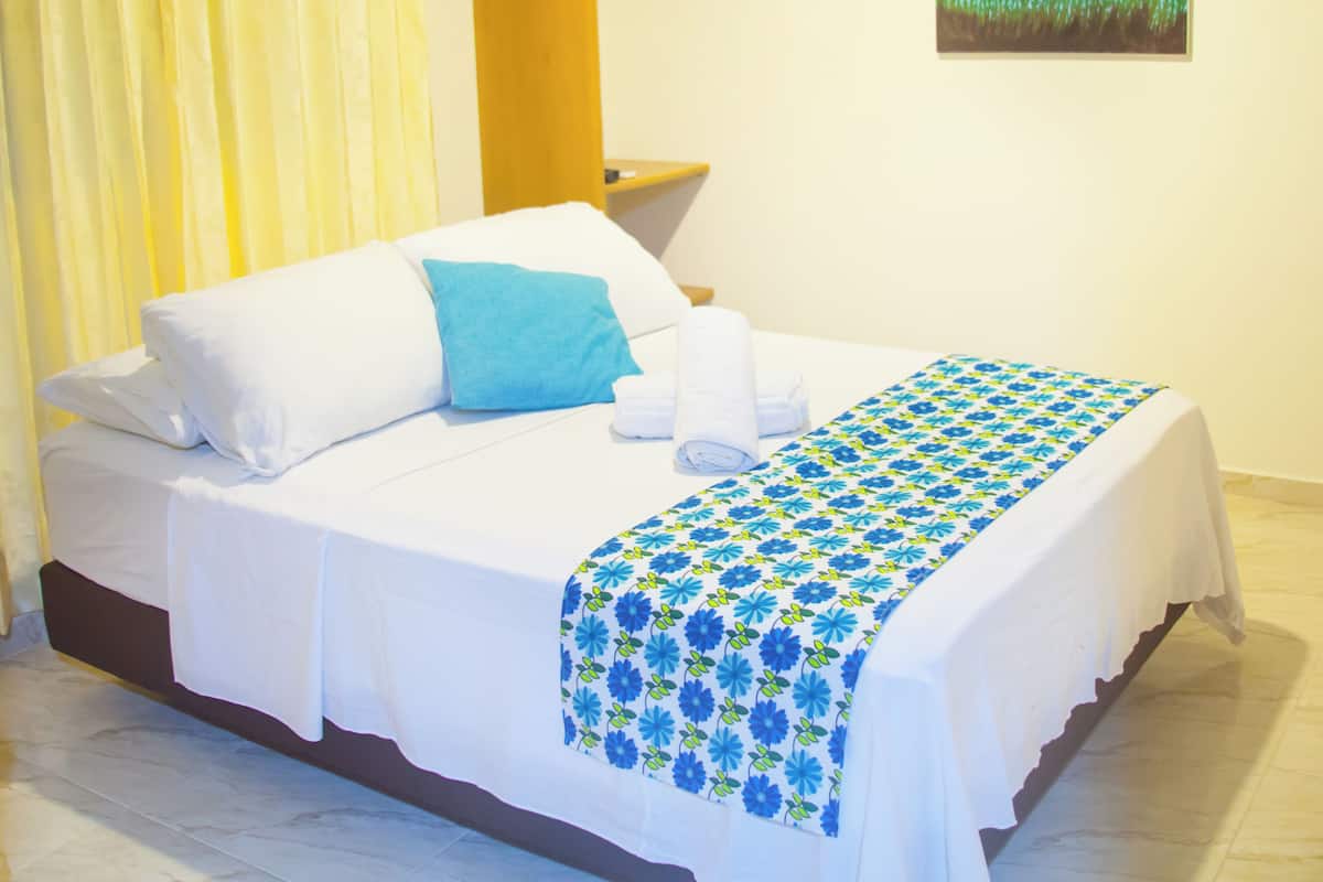 A neatly made bed with white linens and a blue floral bedspread, a yellow curtain, a wooden headboard, and a painting of flowers on the wall.