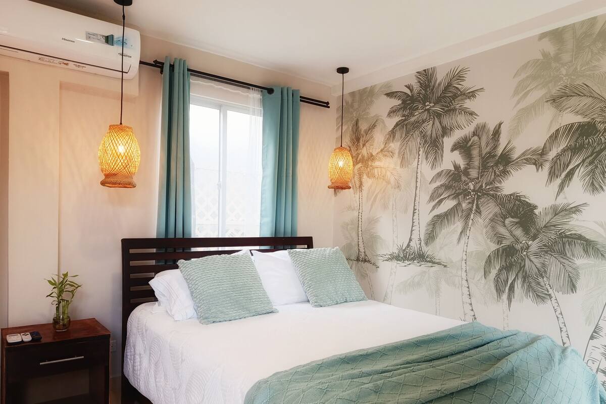 Un dormitorio con cama, mesita de noche, aire acondicionado de pared, ventana con cortinas y un mural de palmera en la pared.