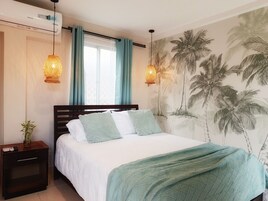 Un dormitorio con cama, mesita de noche, aire acondicionado de pared, ventana con cortinas y un mural de palmera en la pared.