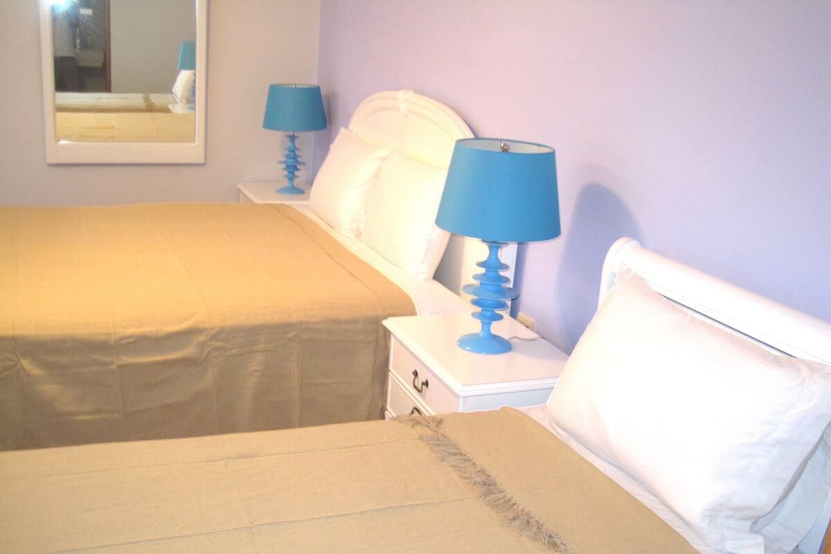 A hotel room with two beds, each with a white headboard and a blue lamp on a bedside table.