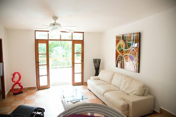 A living room with a white sofa, a glass coffee table, a ceiling fan, and a large abstract painting on the wall.