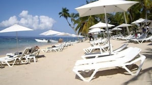A beach with white lounge chairs and umbrellas, palm trees, and a clear sky.