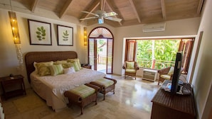 A spacious bedroom with a large bed, a ceiling fan, and a balcony with a view of lush greenery.