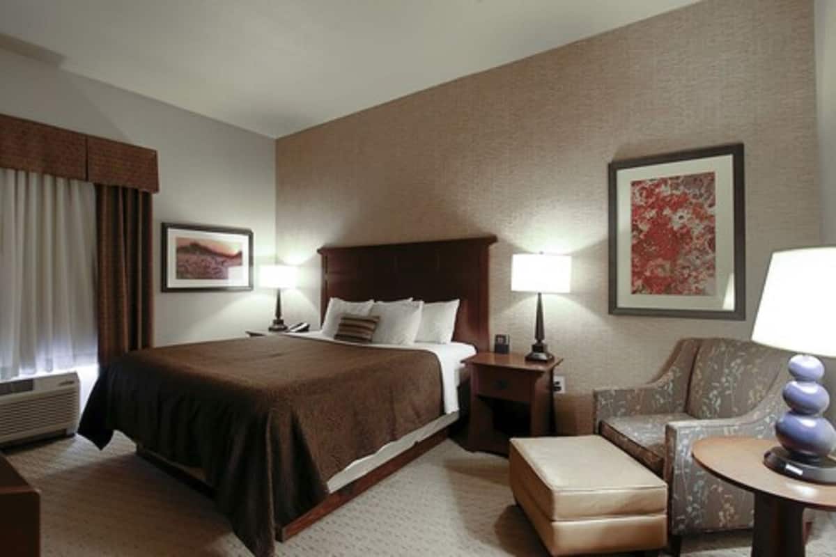 A hotel room with a large bed, two bedside tables with lamps, a chair, an ottoman, and a large painting on the wall.
