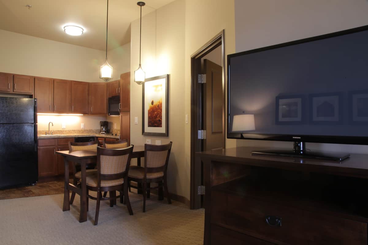 A modern kitchen with wooden cabinets, a dining table with chairs, a refrigerator, and a flat-screen TV on a wooden stand.