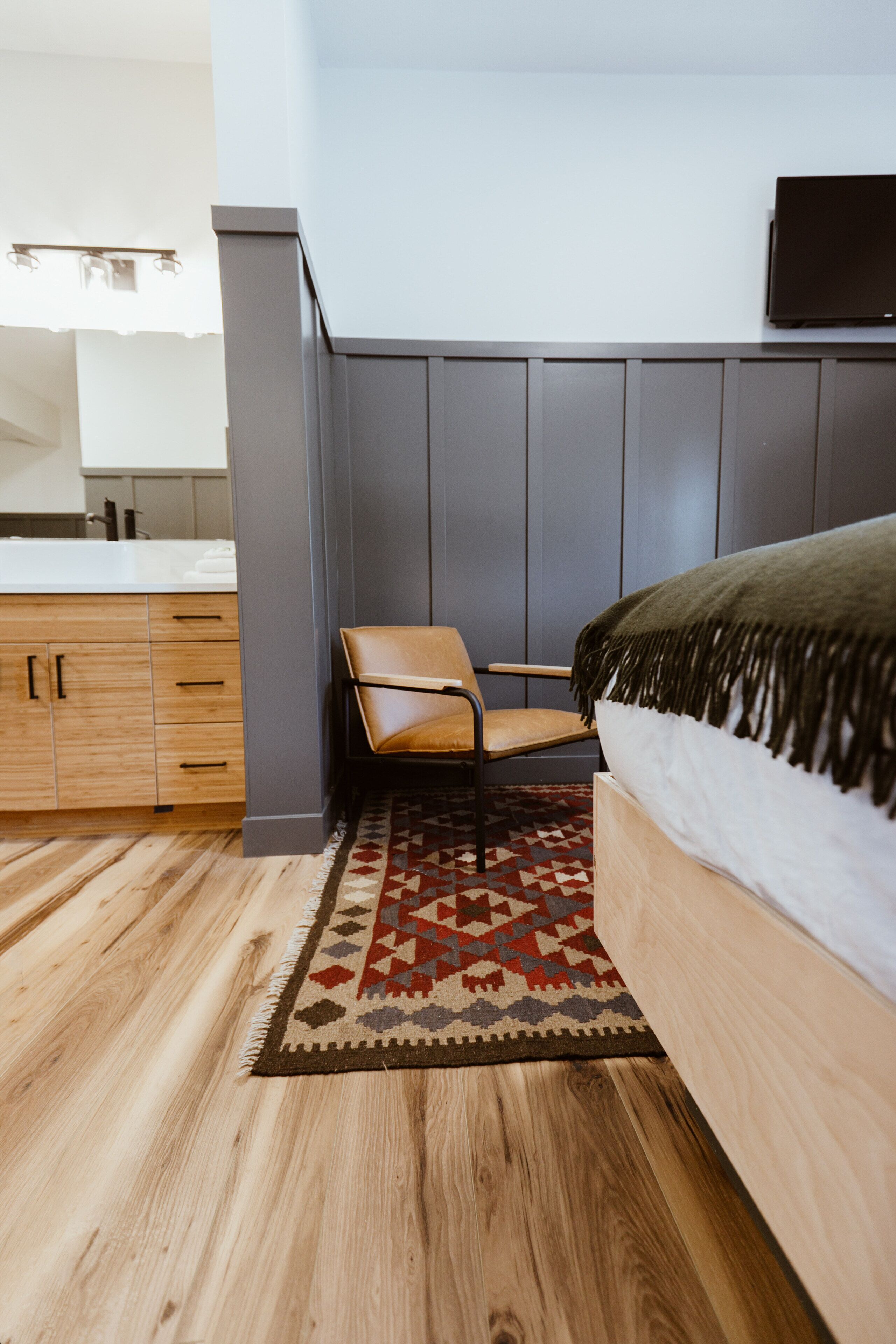 A bedroom with a bed, a chair, a TV mounted on the wall, and a kitchen area with cabinets and a countertop.