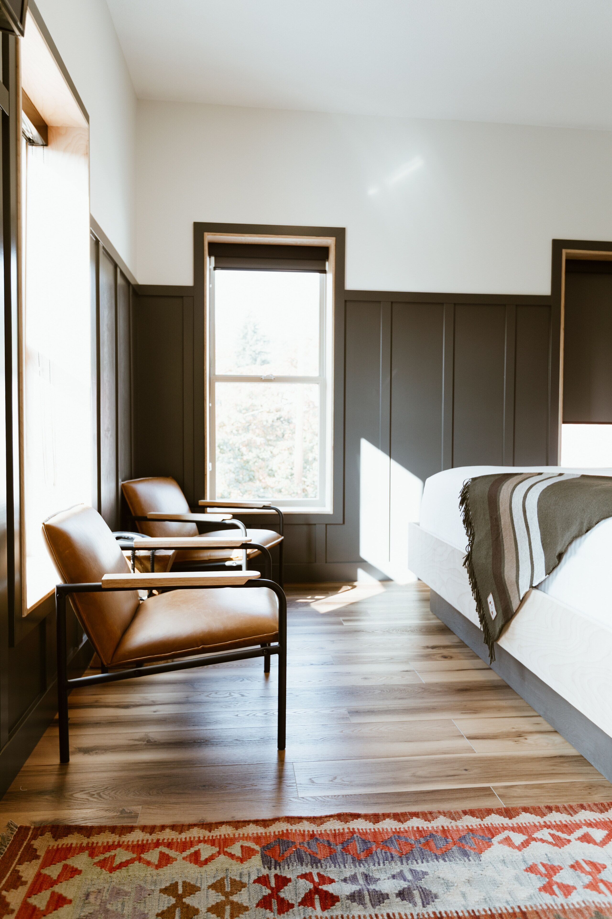 A modern bedroom with a large window, a bed with a striped blanket, two leather chairs, and a patterned rug.