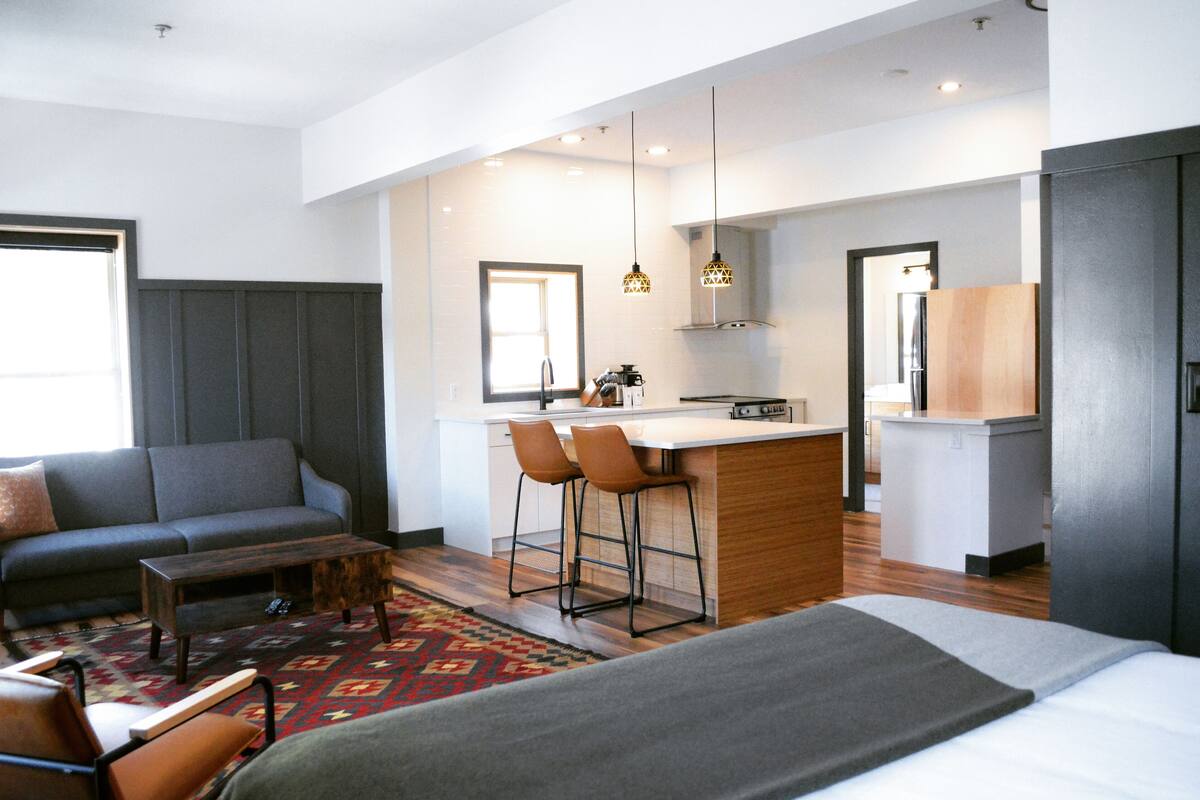 Ett modernt hotellrum med en soffa, ett litet soffbord, en köksdel med en barstol och en stor säng.
