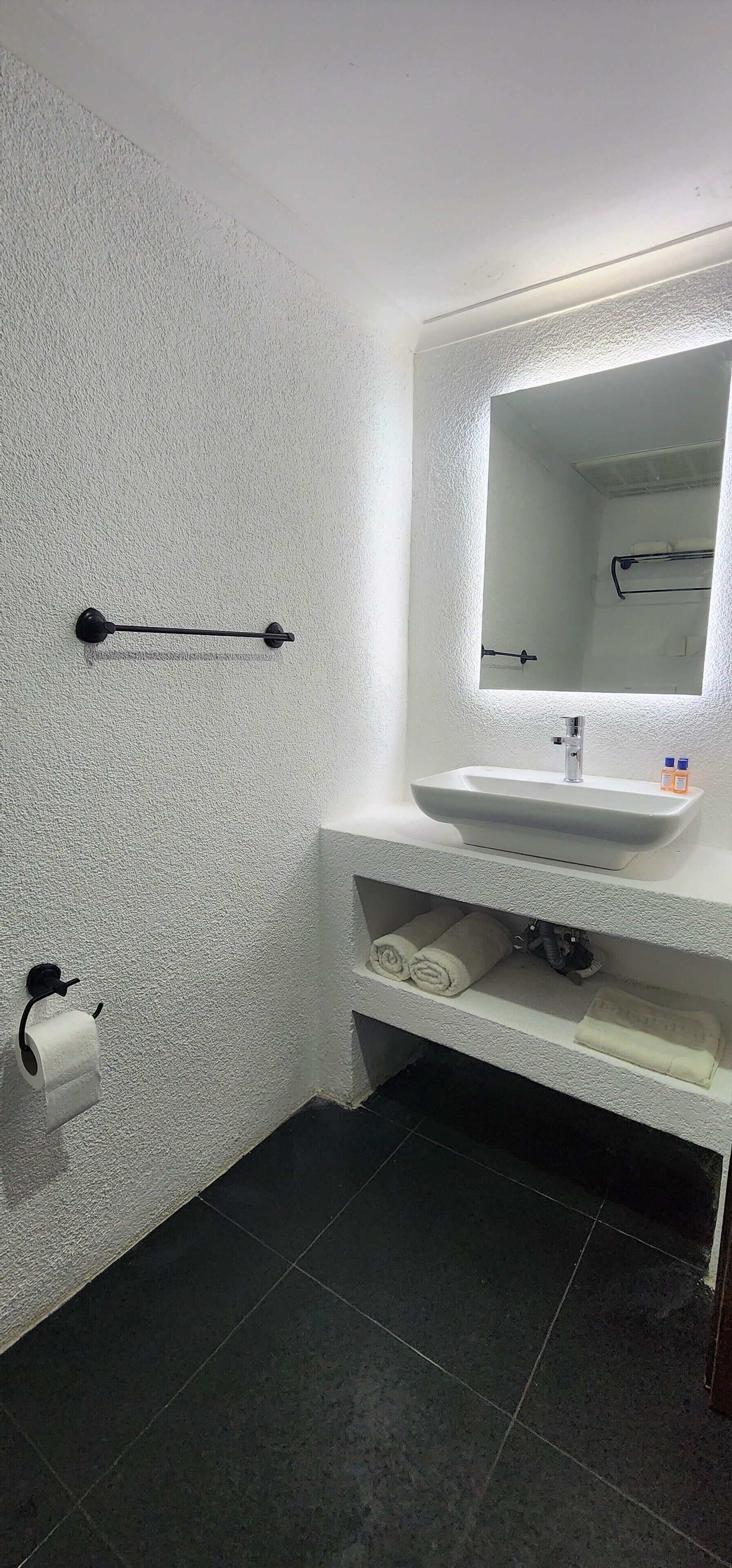 A bathroom with a white textured wall, a black towel rack, a white sink with a mirror, and dark tiled flooring.