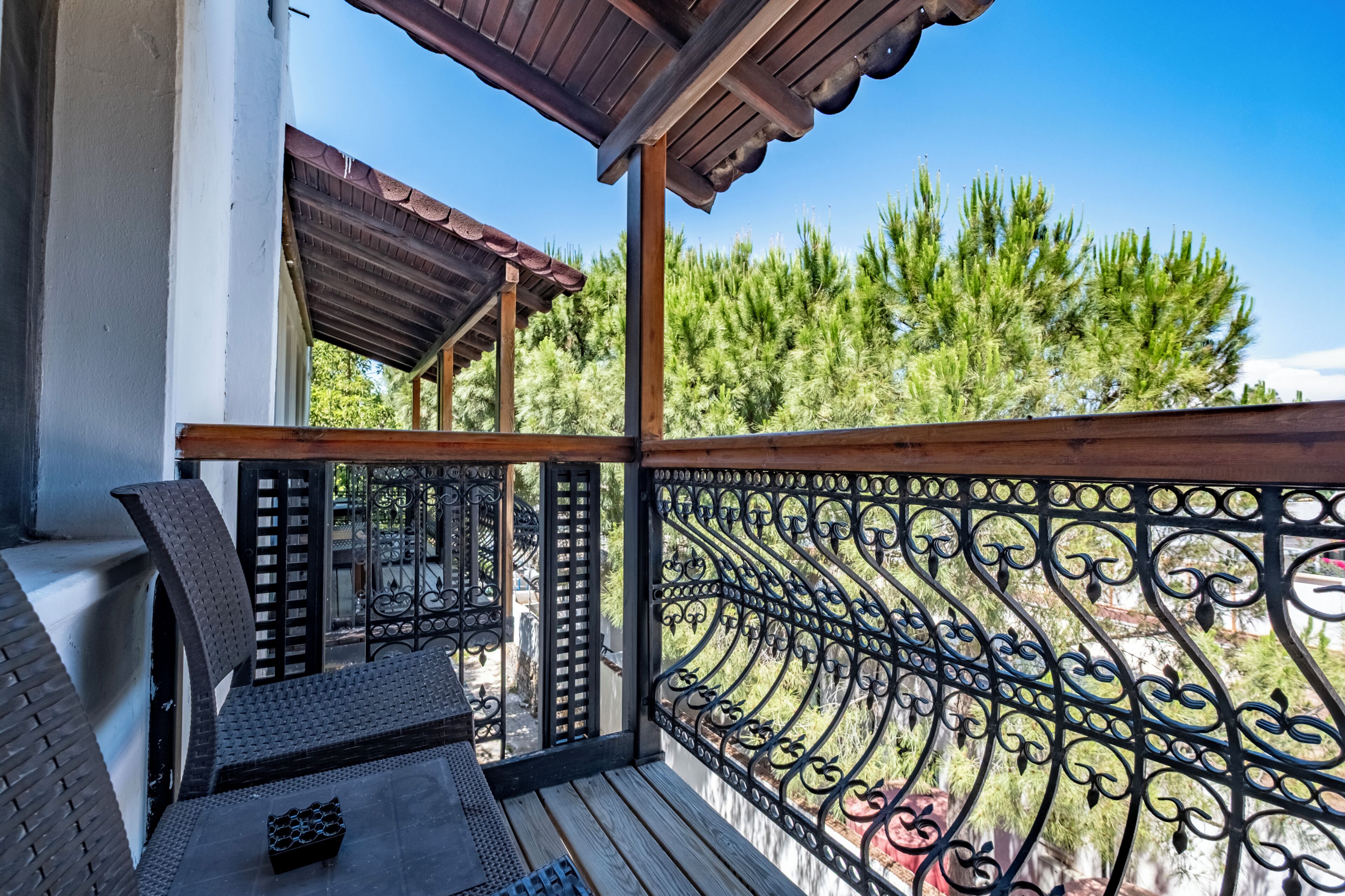A balcony with a wooden railing, a seating area with a bench and a table, and a view of trees and a building in the background.