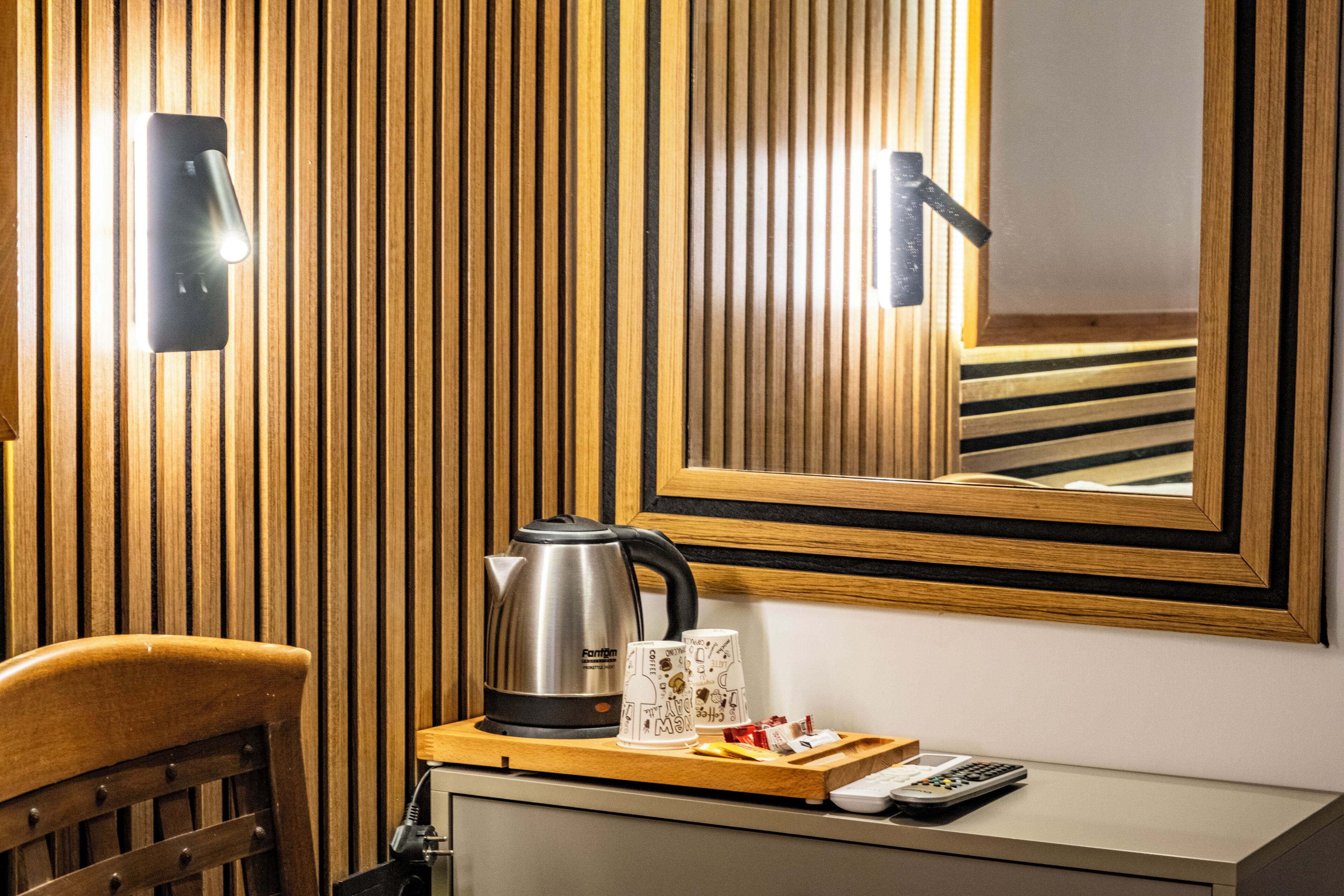 A wooden-paneled wall with a kettle, two mugs, a tray with items, a remote control, and a mirror reflecting the room.