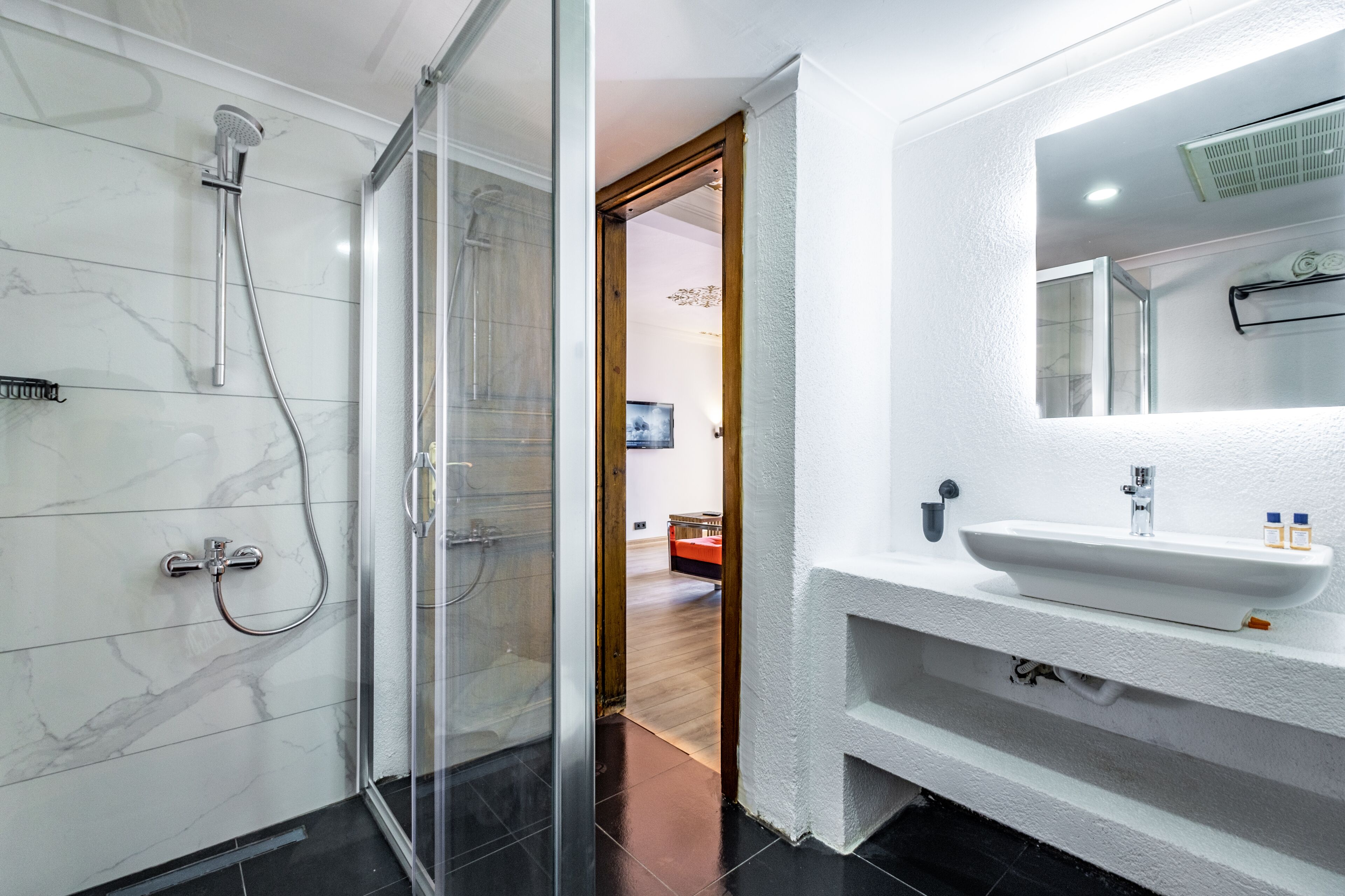 Un baño moderno con cabina de ducha de vidrio, un lavabo blanco con espejo y una puerta de madera que da a otra habitación.