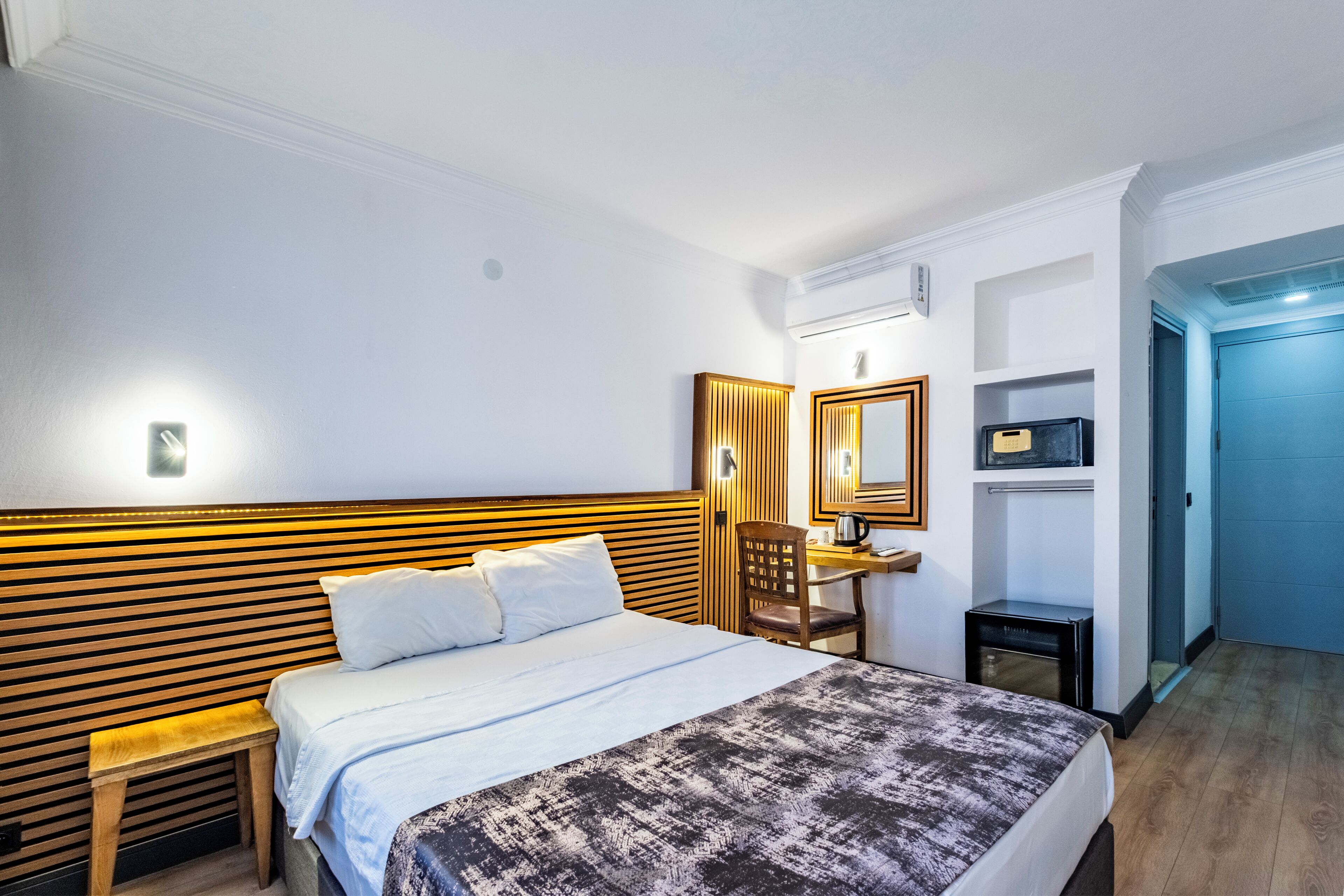 A hotel room with a bed, a desk, a chair, a mirror, and a blue door.