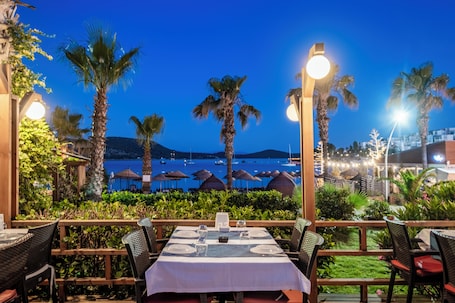 A restaurant with outdoor seating overlooking a body of water, palm trees, and a mountainous backdrop.