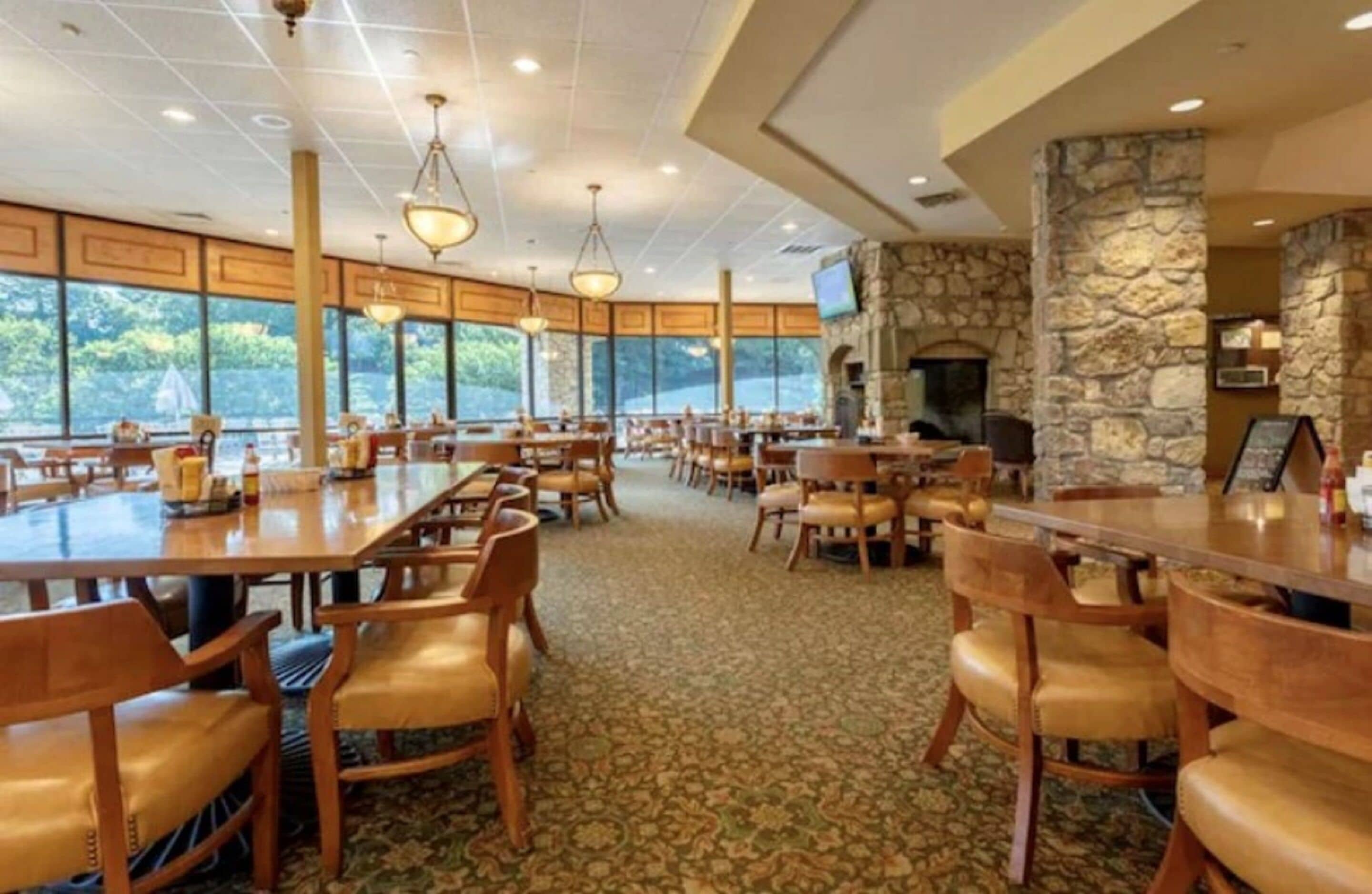 A spacious dining area with multiple tables and chairs, large windows, and a stone fireplace.
