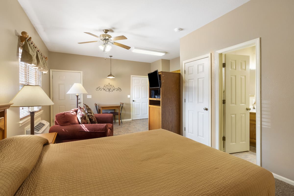 A hotel room with a large bed, a desk, a chair, a ceiling fan, and a door leading to another room.