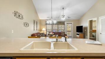 A kitchen with a large island, double sink, and a ceiling fan. There is a dining area with a couch and a TV in the background.