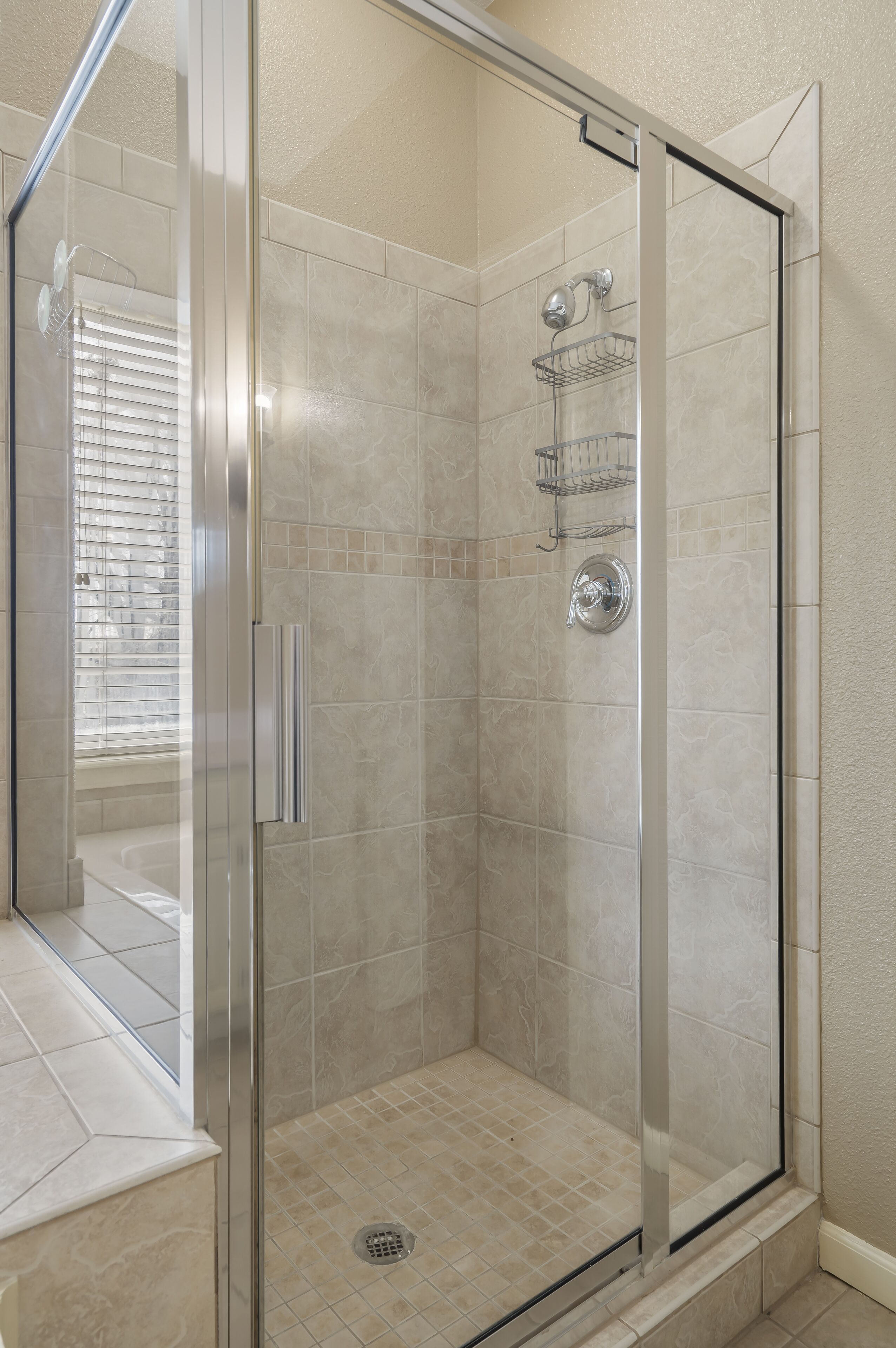 A glass-enclosed shower stall with a tiled floor, a metal shower head, and a wall-mounted shelf.