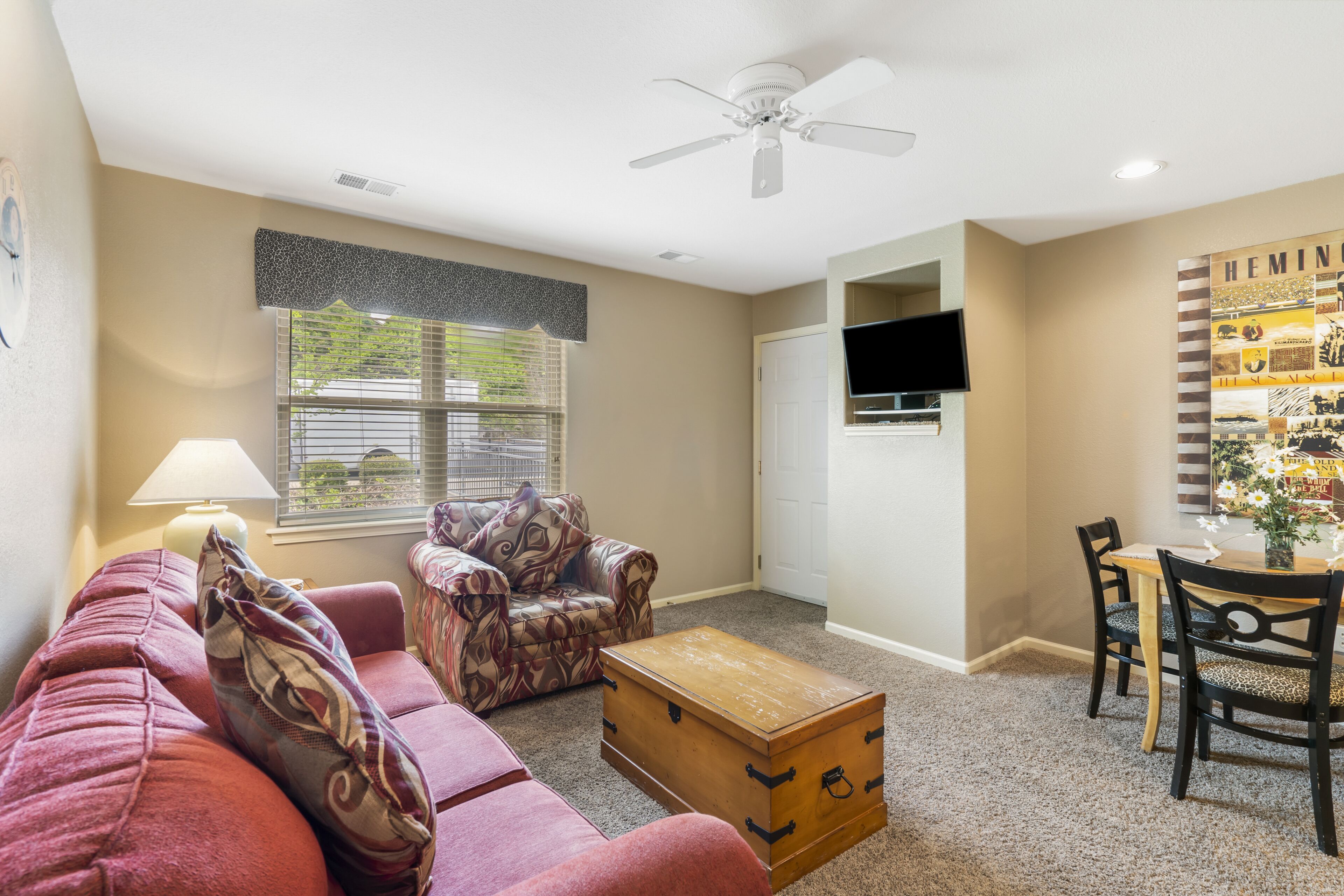 A living room with a sofa, a patterned armchair, a wooden chest, a dining table with chairs, a ceiling fan, and a wall-mounted TV.