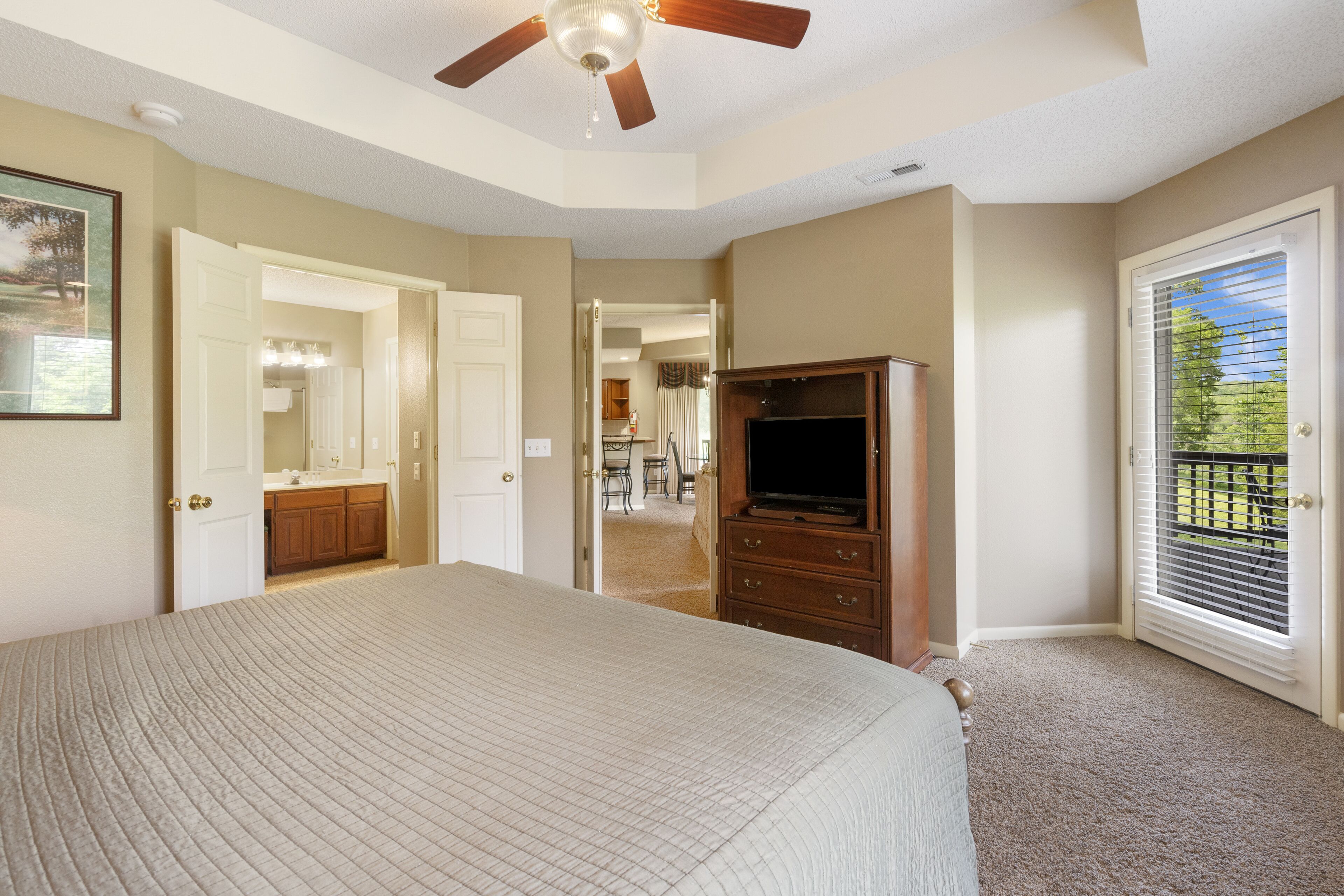 A bedroom with a large bed, a ceiling fan, a dresser, and a TV. There is a door leading to a balcony with a view of the outdoors.
