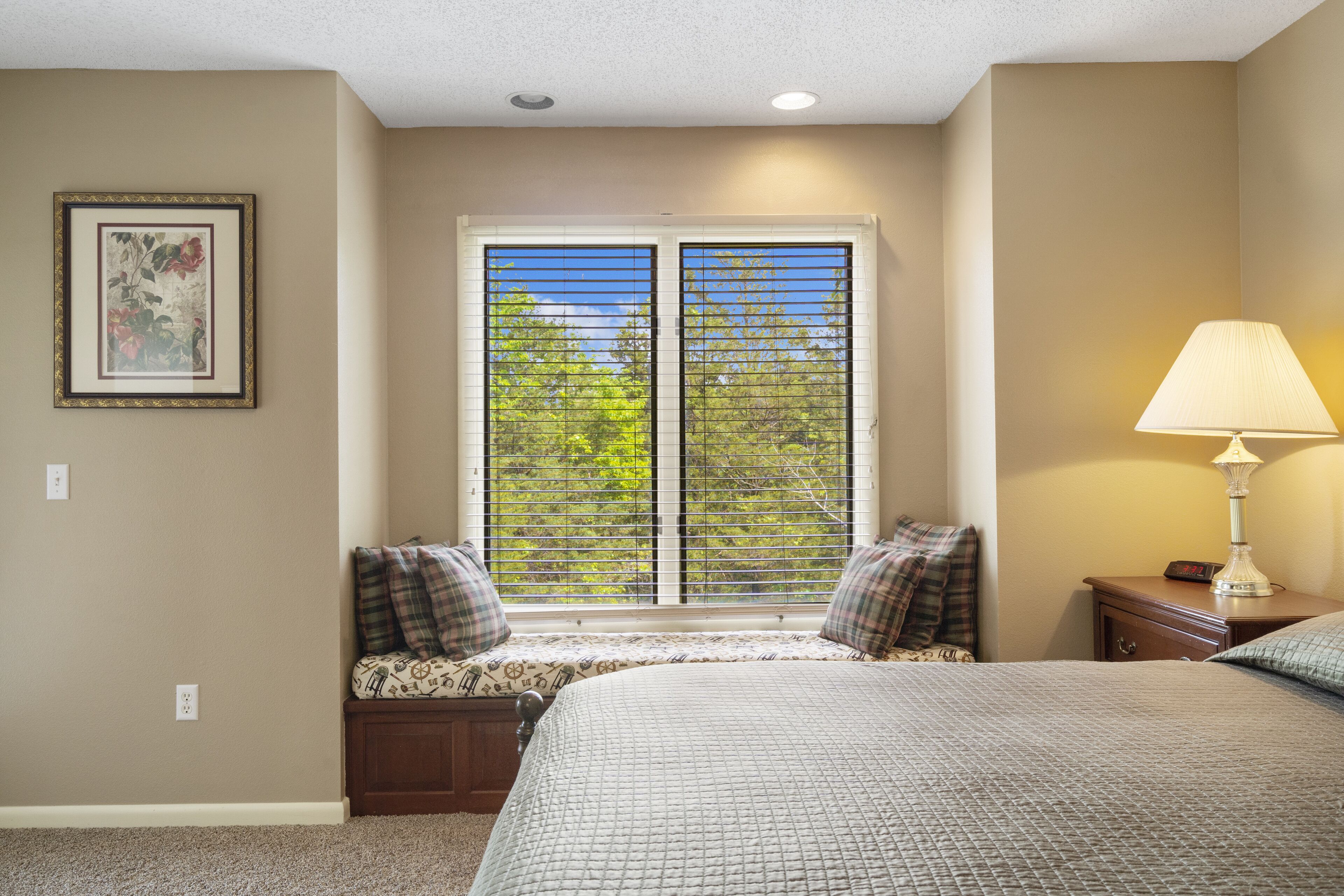A bedroom with a large bed, a window with blinds, a bedside table with a lamp, and a framed picture on the wall.