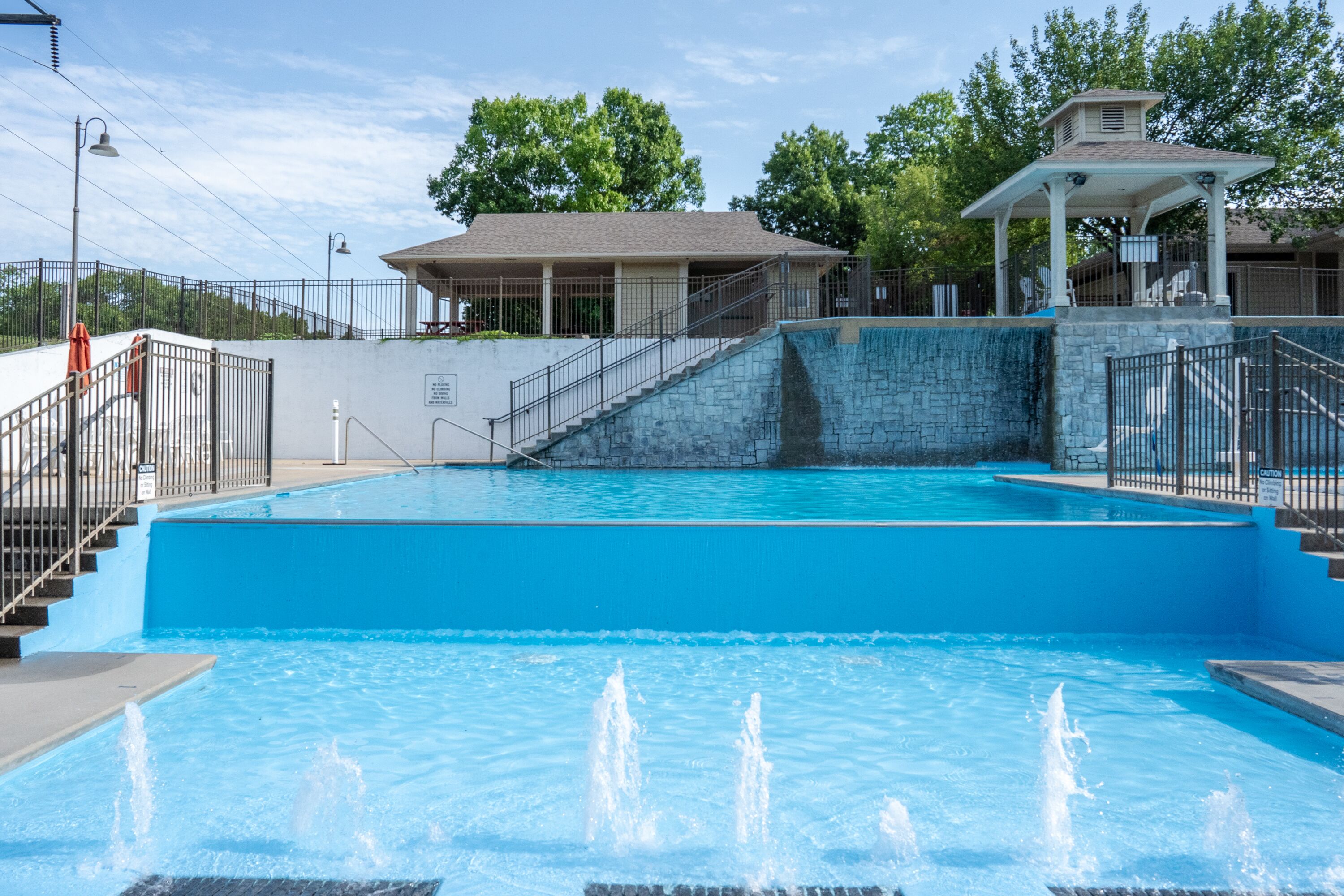 Une piscine bordée d’un mur en pierre, un espace de repos couvert et un escalier menant à un bâtiment.