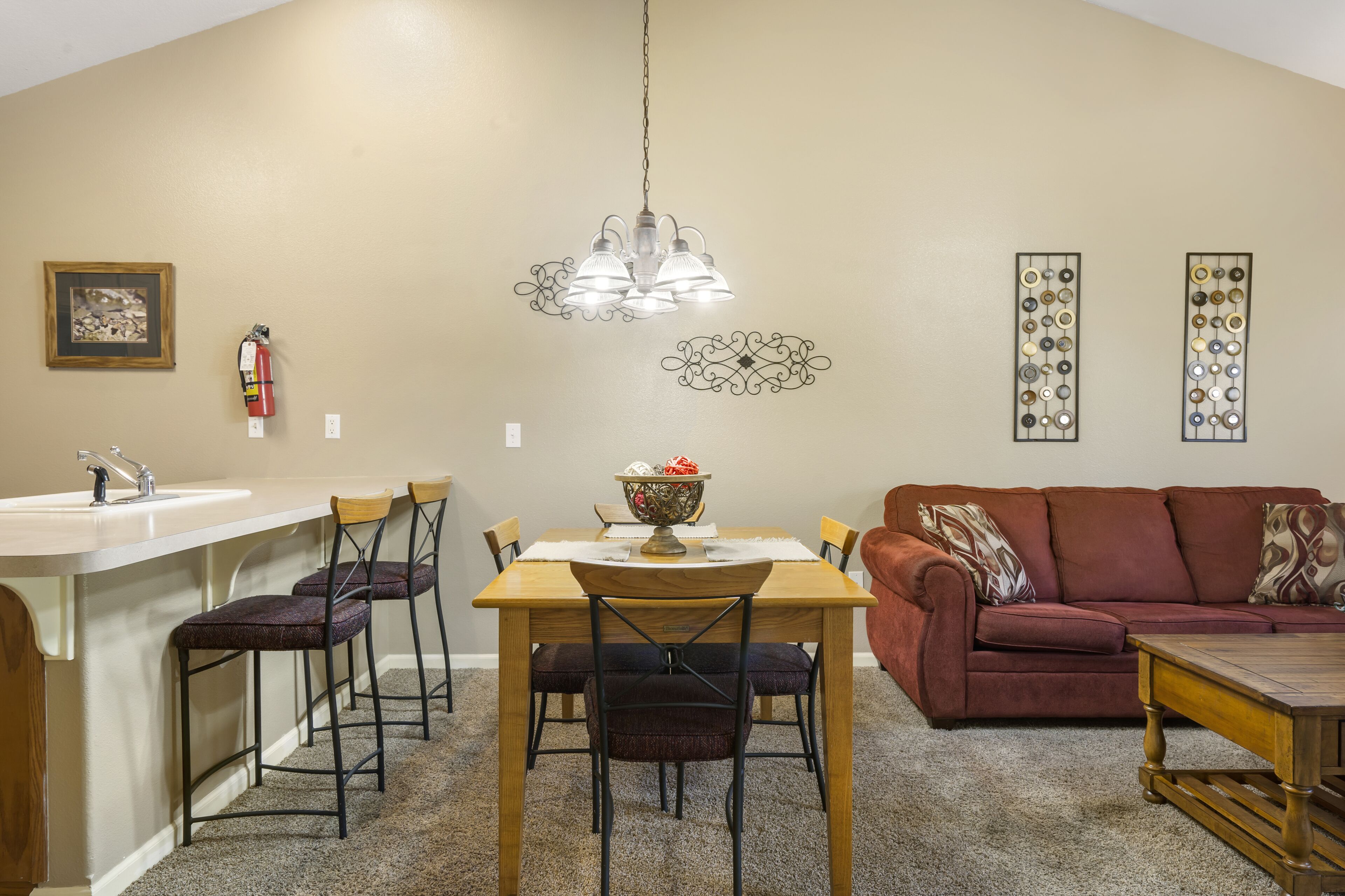 A dining area with a wooden table, four chairs, a chandelier, a red couch, and a small kitchen area with a sink and a fire extinguisher.
