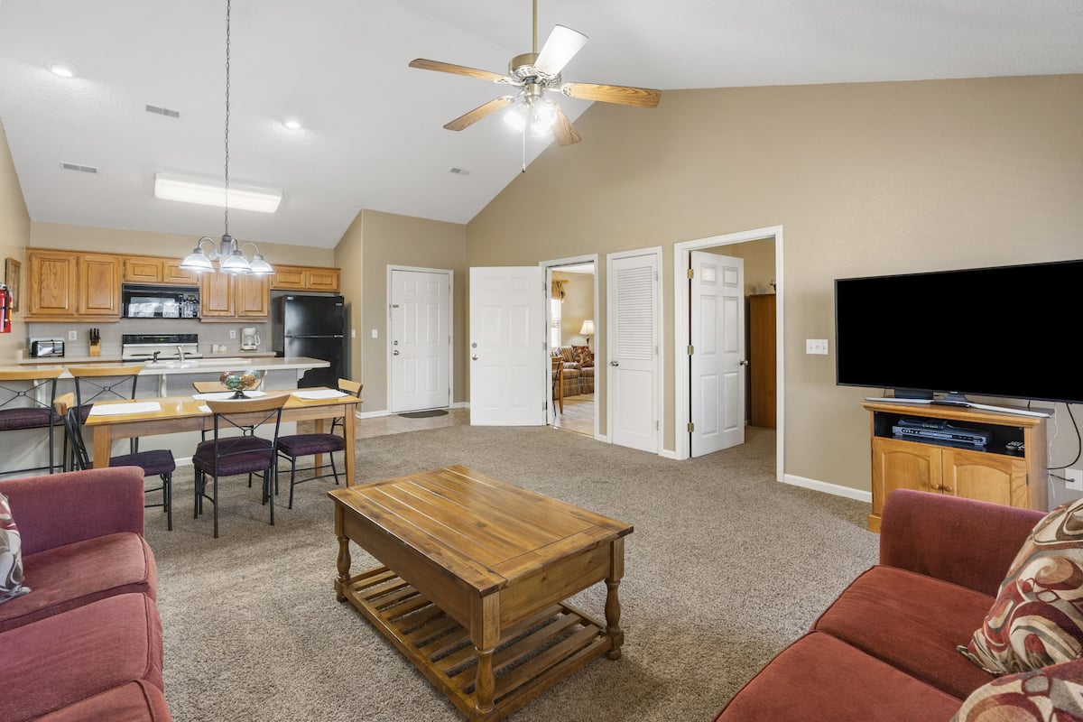 A spacious living area with a wooden dining table, red sofas, a ceiling fan, and a television on a wooden cabinet.