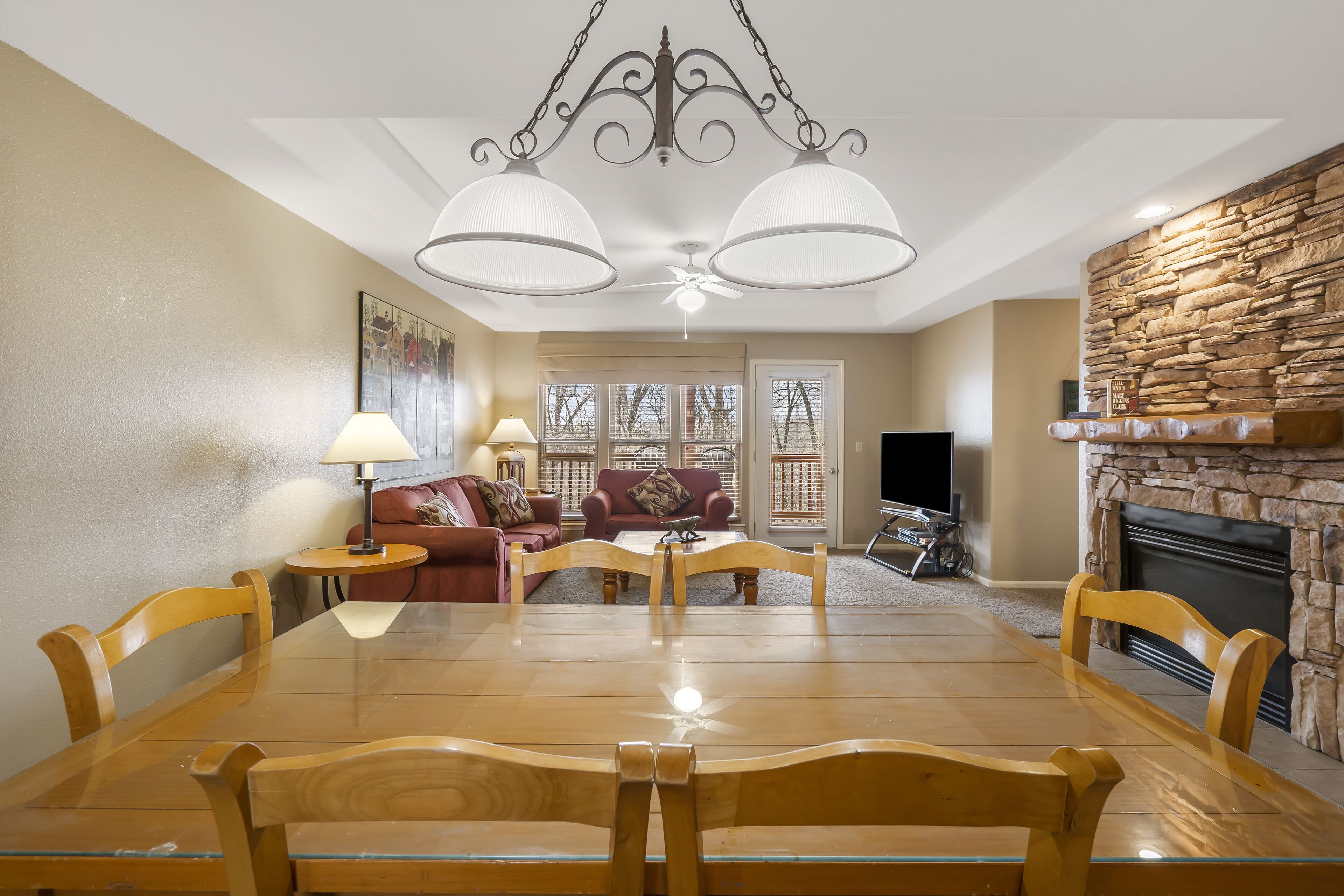 A dining area with a wooden table and chairs, a chandelier, a stone fireplace, and a living room with a sofa and TV.