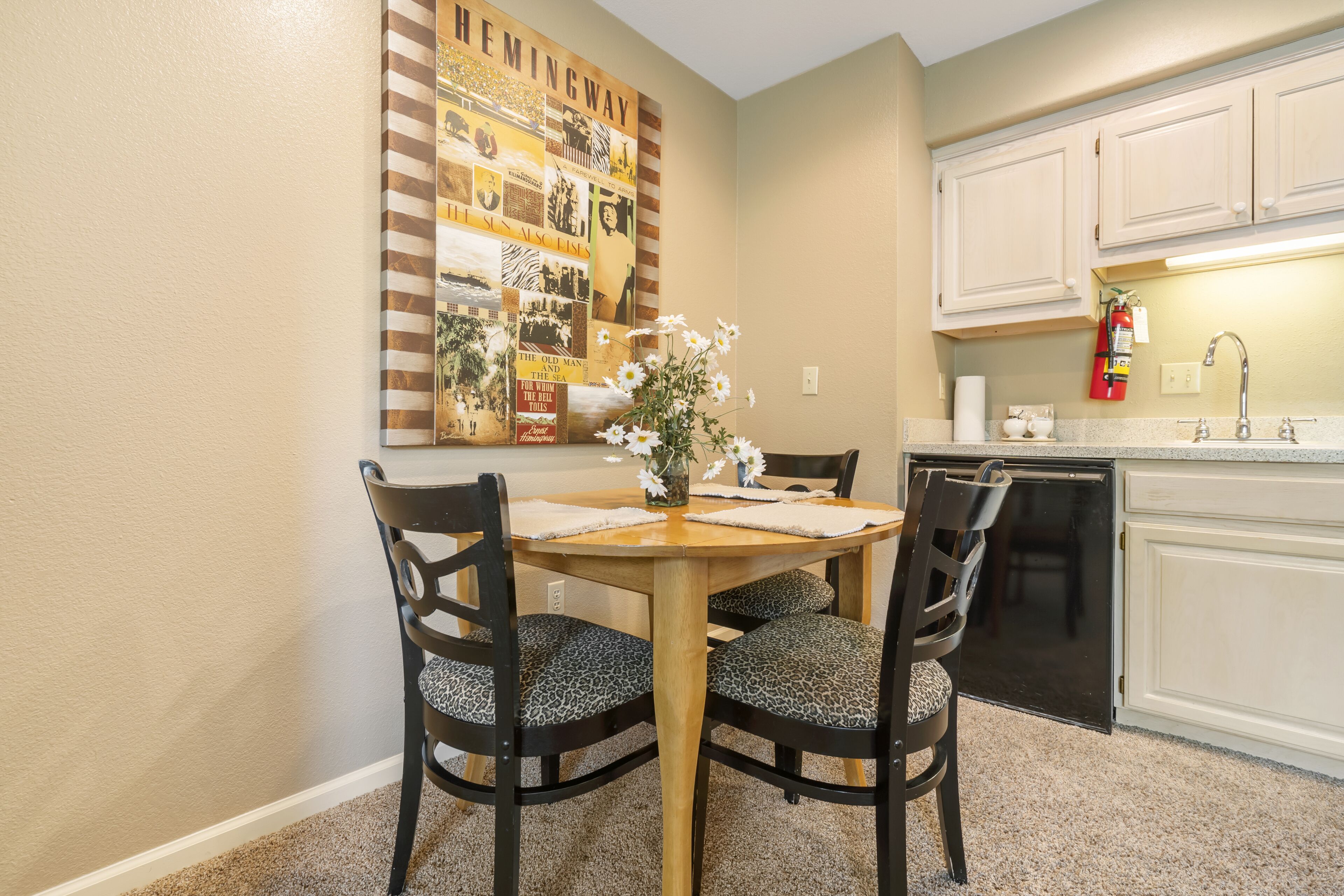 A dining area with a wooden table and chairs, a wall-mounted collage, and a kitchen with white cabinets and a dishwasher.