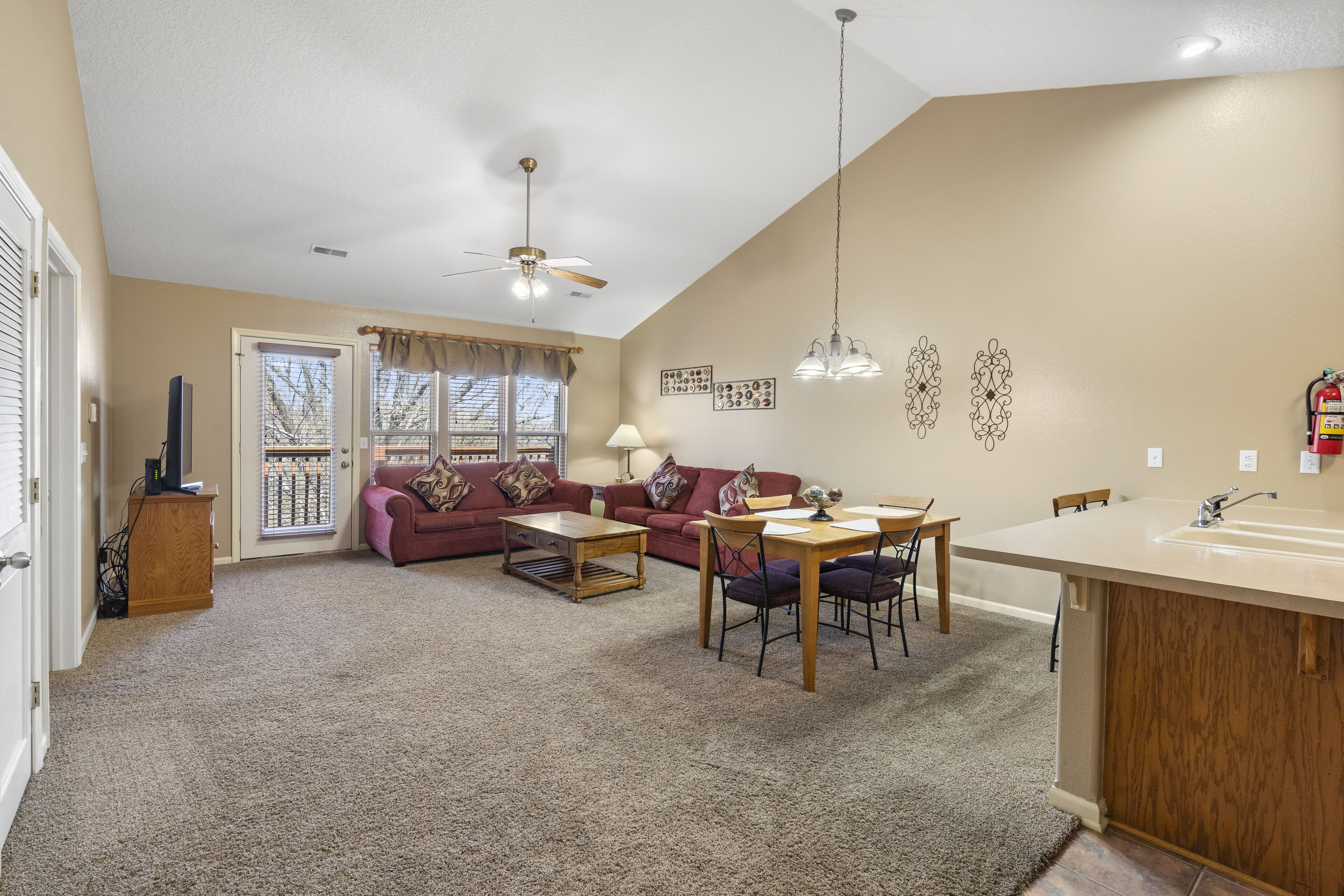 A living room with a sofa, coffee table, and dining table. There is a kitchen area with a sink and a ceiling fan.