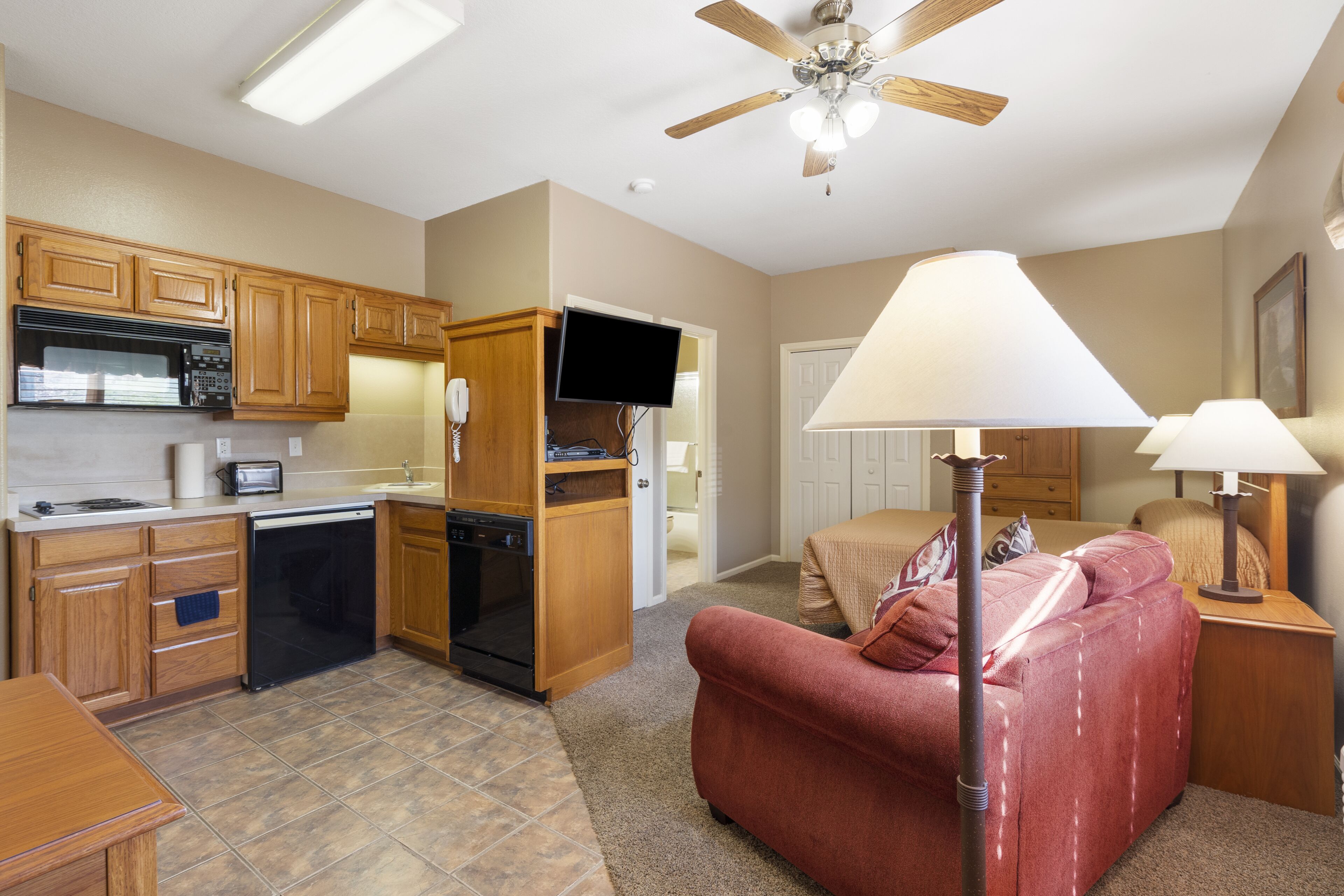 A compact kitchen with wooden cabinets, a dishwasher, and a microwave. There is a red sofa and a lamp in the living area.
