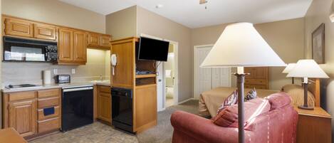 A compact kitchen with wooden cabinets, a dishwasher, and a microwave. There is a red sofa and a lamp in the living area.