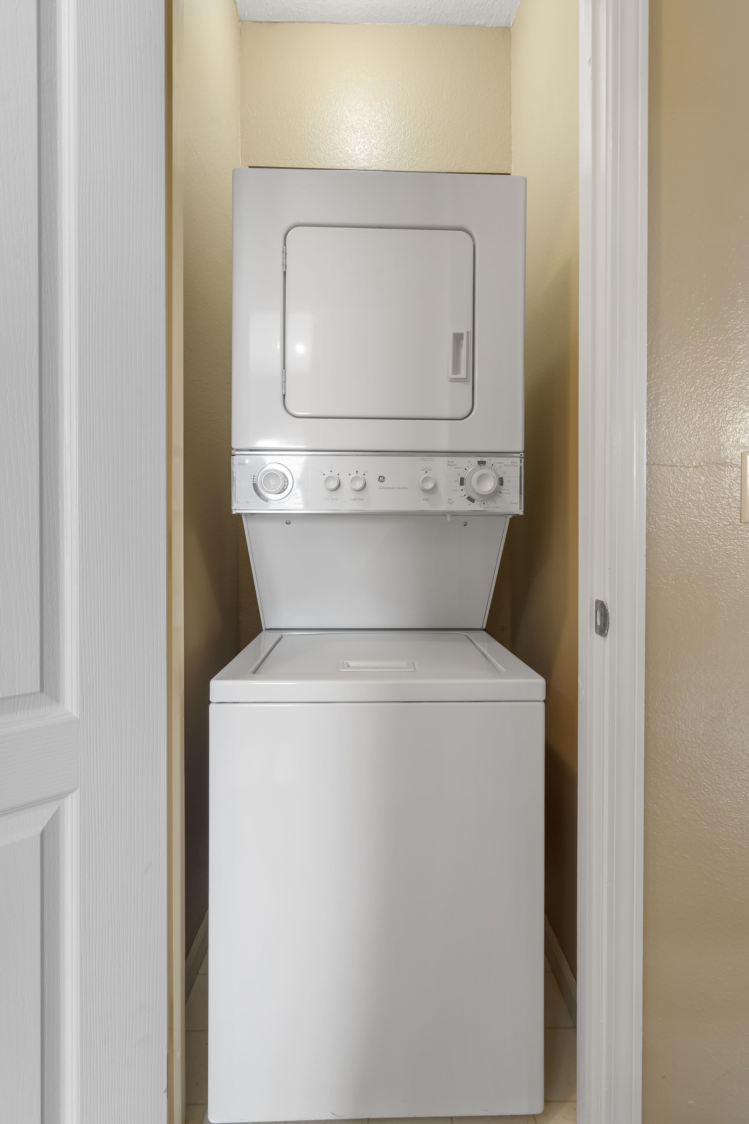 A compact, white stacked washer and dryer unit in a narrow hallway with beige walls.