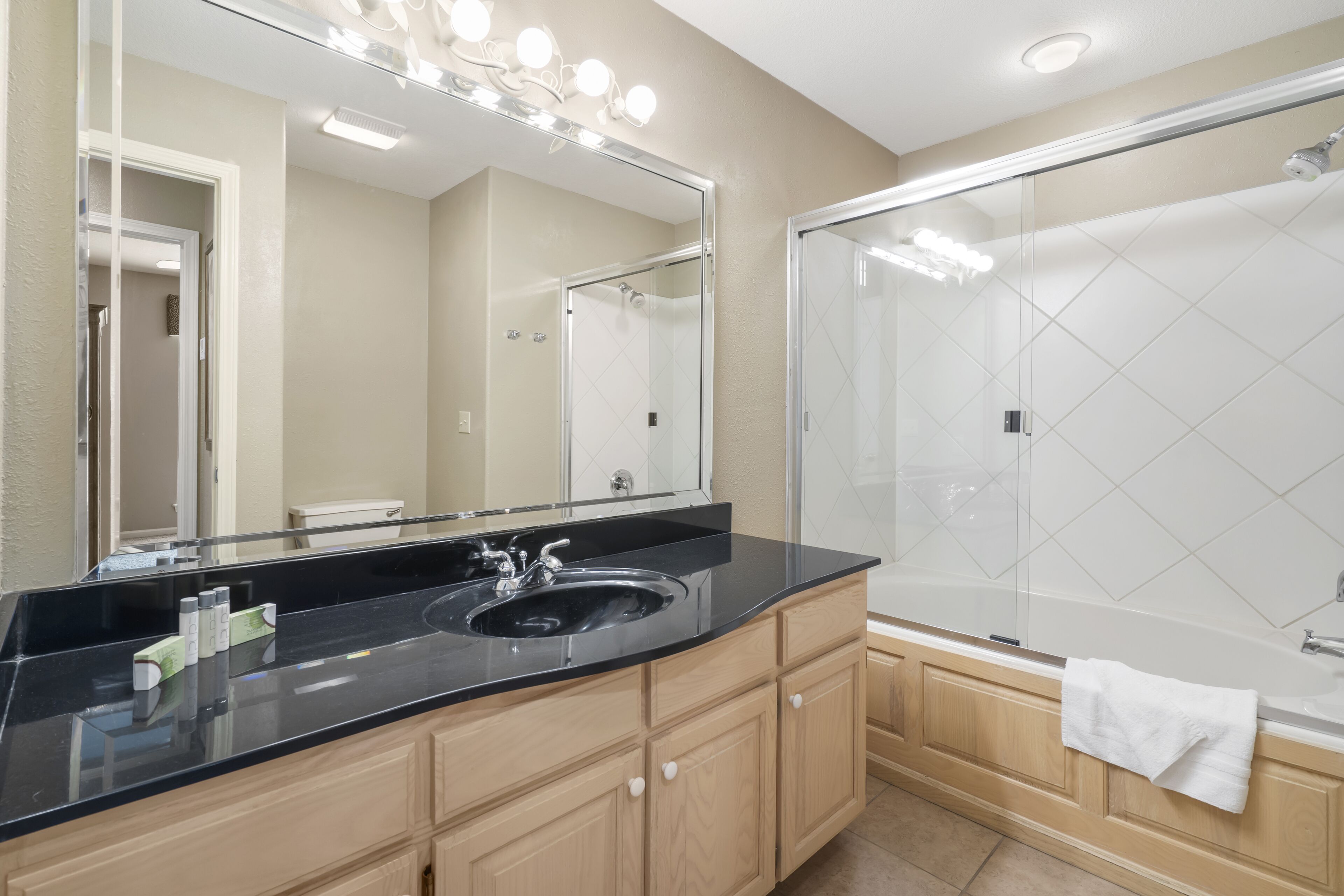 A modern bathroom with a black countertop sink, wooden cabinets, a large mirror with lights, a glass shower enclosure, and a white towel hanging on the bathtub.