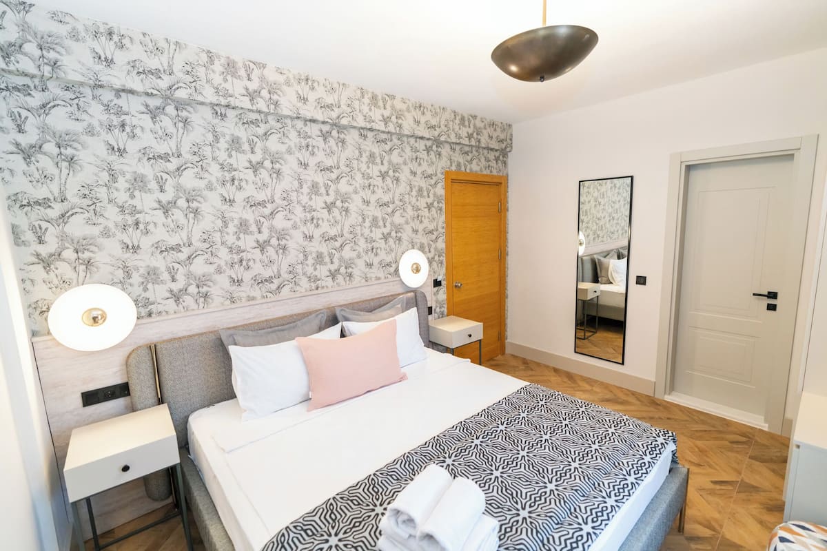 Un dormitorio ordenado con papel tapiz estampado, una cama con colcha geométrica, lámparas de noche, una mesita de noche y un espejo en la pared.