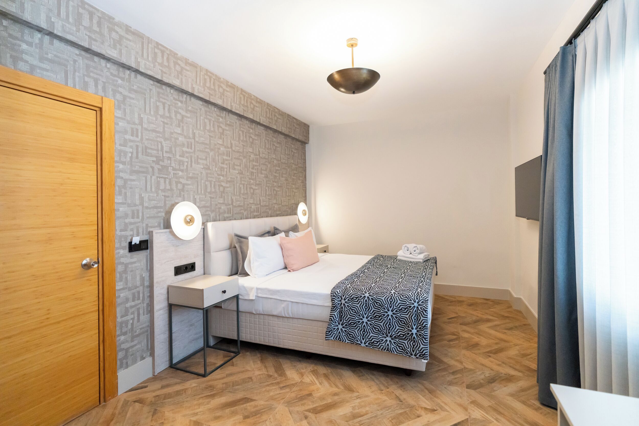 A modern bedroom with a wooden door, a bed with patterned bedding, a bedside table, and a ceiling light.