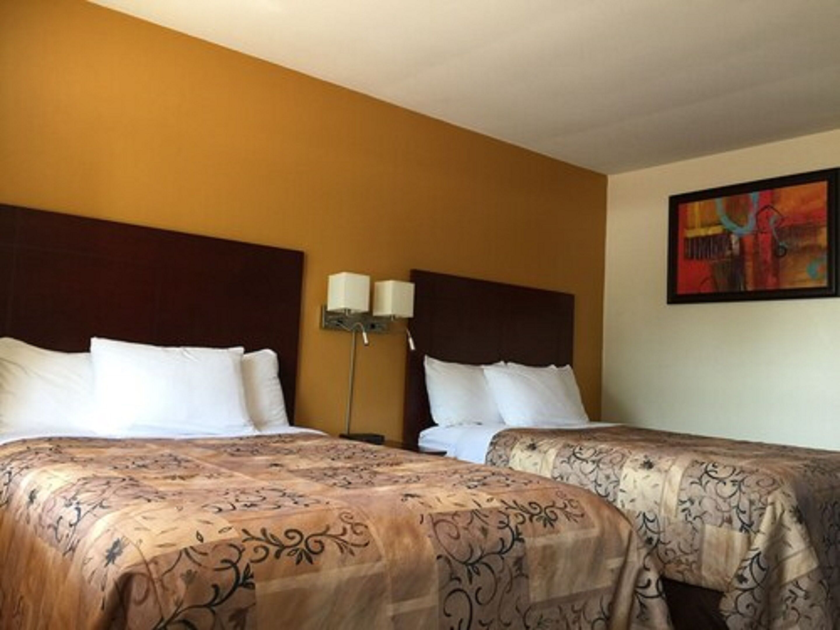 Una camera d'albergo con due letti, entrambi con lenzuola bianche e una coperta a motivi decorativi. Tra i letti c'è una lampada a parete, e sopra il letto a destra c'è un quadro.