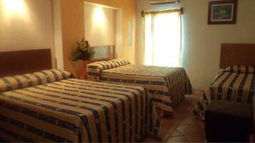 A hotel room with three beds, a potted plant, and a window with curtains.