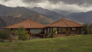 Un belvédère doté d’un toit en tuiles et d’une table, entouré d’une pelouse et avec des montagnes en arrière-plan.
