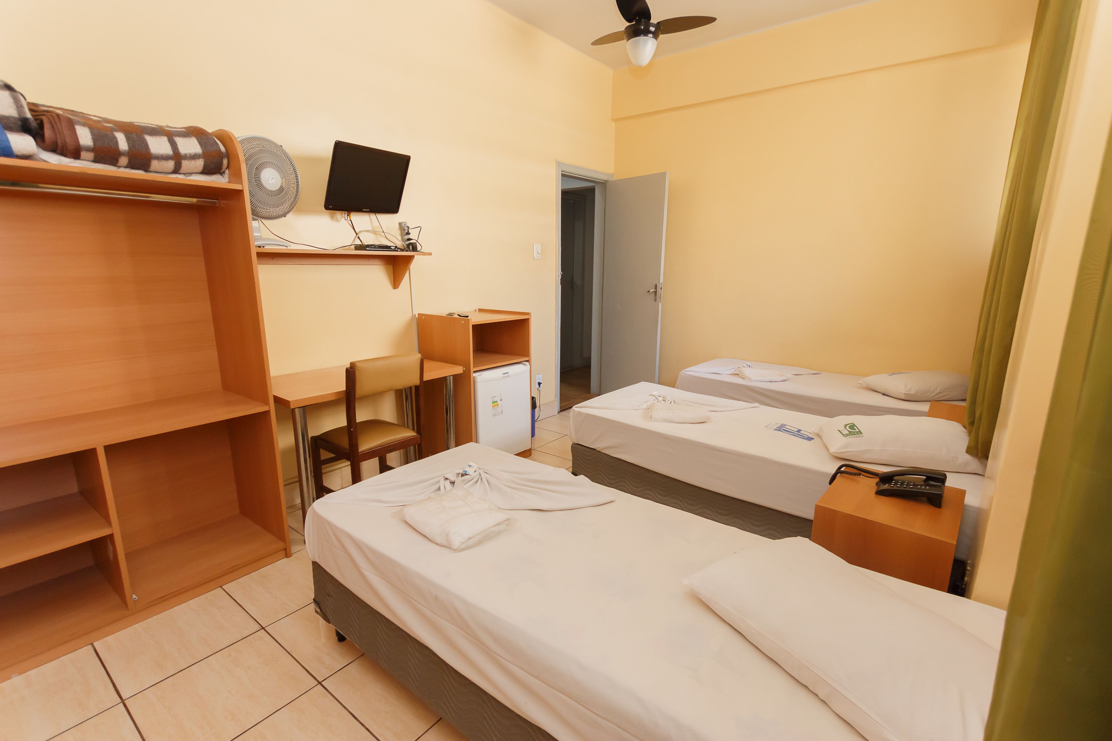 Quarto de hotel com duas camas de solteiro, uma escrivaninha com cadeira, um frigorífico pequeno e uma televisão na parede.