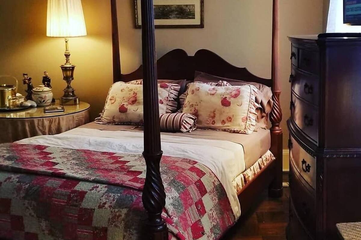 Ein Himmelbett mit floraler Bettwäsche, ein Nachttisch mit Lampe und eine Kommode mit Schubladen.