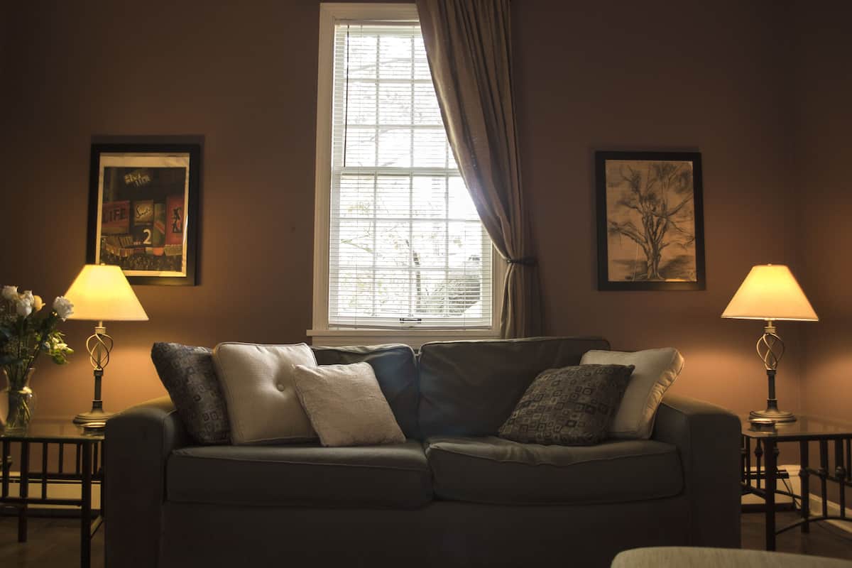 Ein Wohnzimmer mit einer Couch, zwei Lampen, einem Fenster mit Vorhängen und zwei gerahmten Bildern an der Wand.