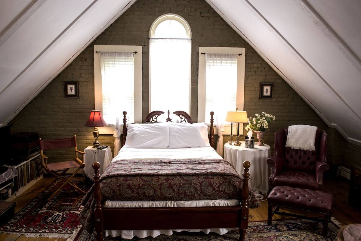 Ein Schlafzimmer mit einem großen Bett, zwei Nachttischen, einem Sessel und einem Fenster mit Vorhängen.