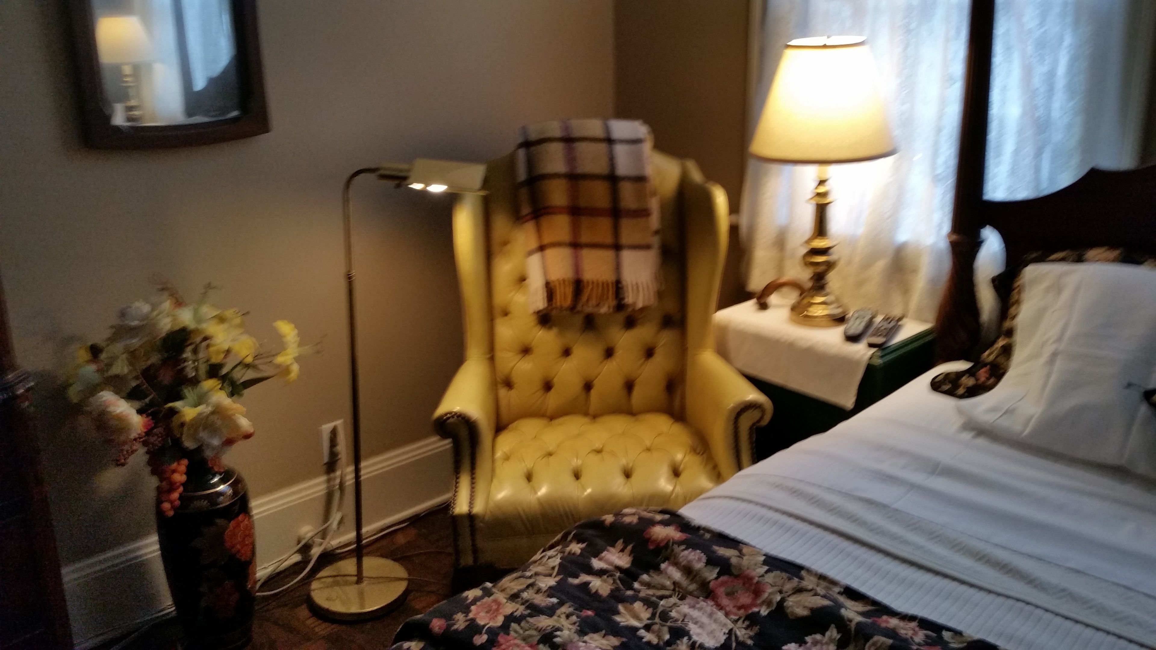 Een hotelkamer met een bed, een gele fauteuil, een nachtkastje met een lamp, een vloerlamp en een bloemstuk.
