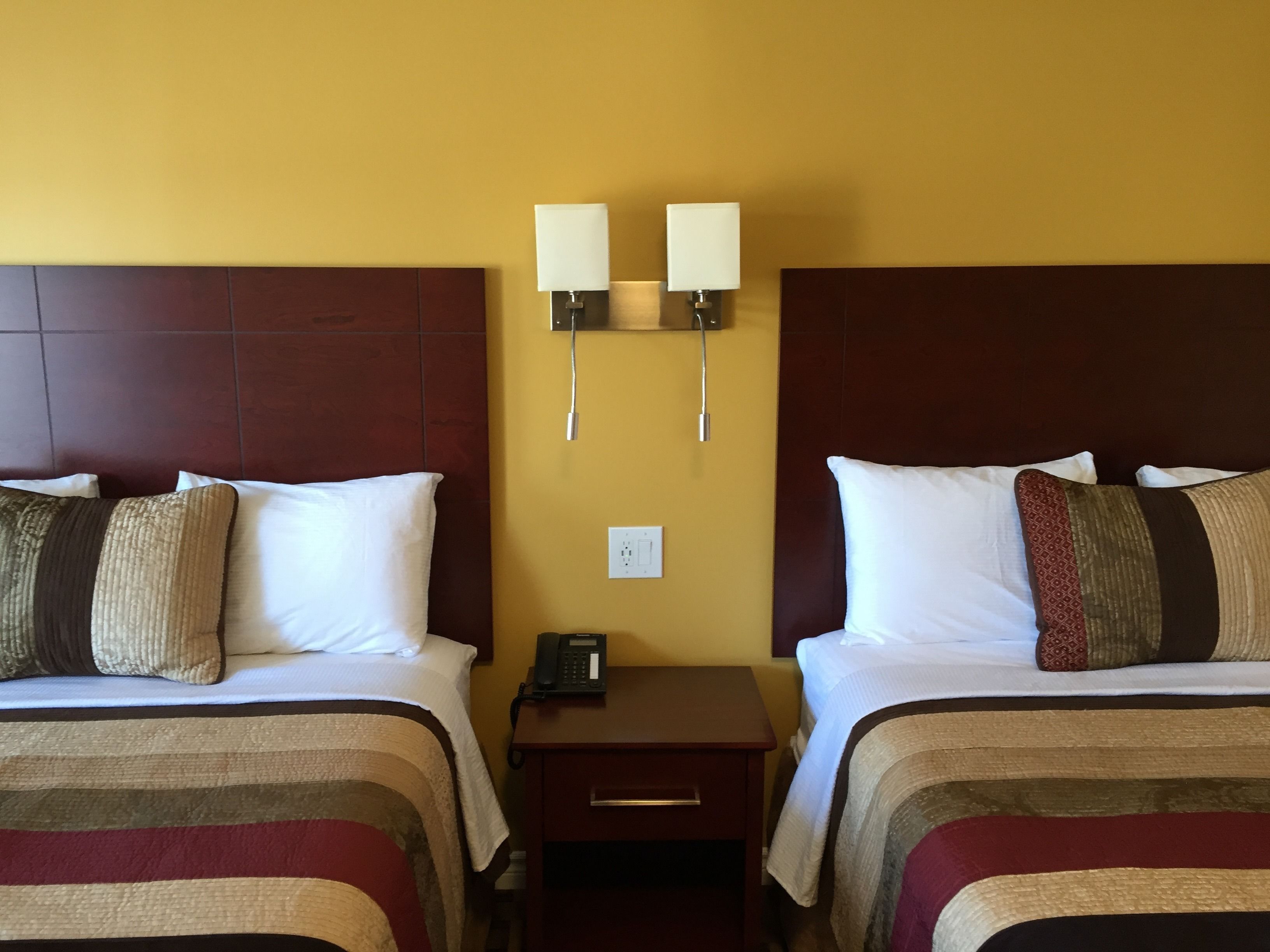 A hotel room with two beds, each with a striped bedspread, a nightstand with a phone, and two wall-mounted lamps.
