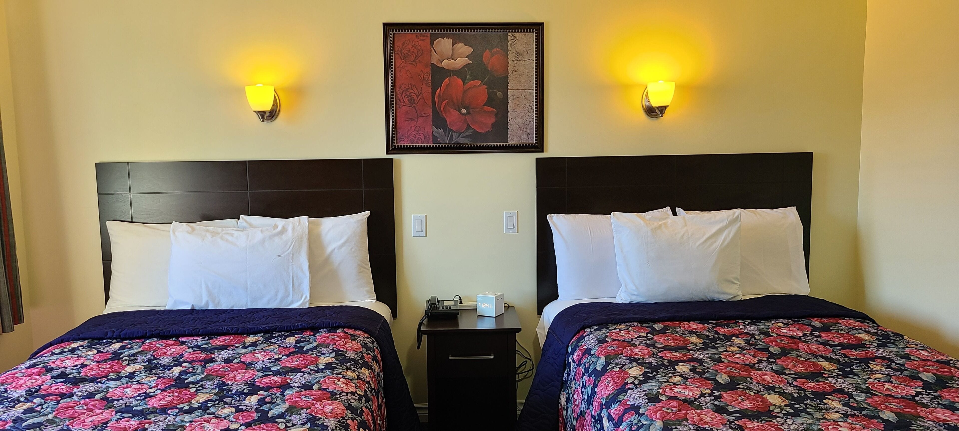 Une chambre d’hôtel avec deux lits, chacun doté d’une tête de lit sombre et de draps blancs. De chaque côté d’un tableau représentant des motifs floraux, on trouve deux appliques murales.
