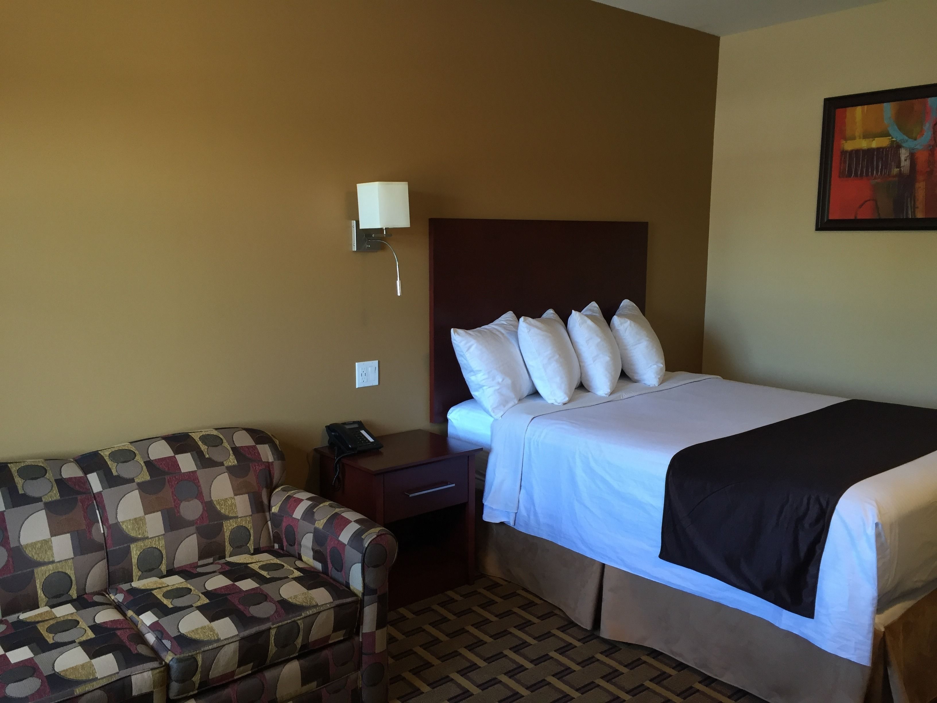 A hotel room with a bed, a patterned armchair, a bedside table with a phone, and a wall-mounted lamp.