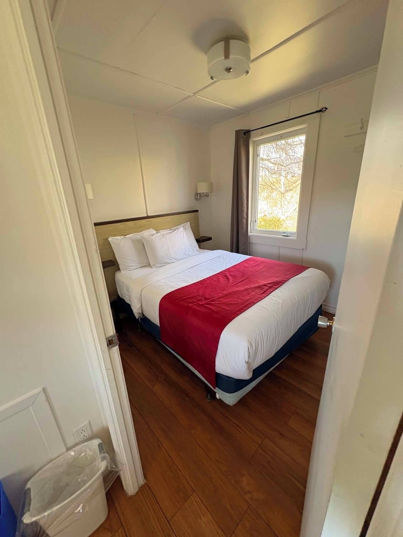 Una cama individual con ropa de cama blanca y roja, suelo de madera, una ventana con cortinas y una lámpara en el techo.